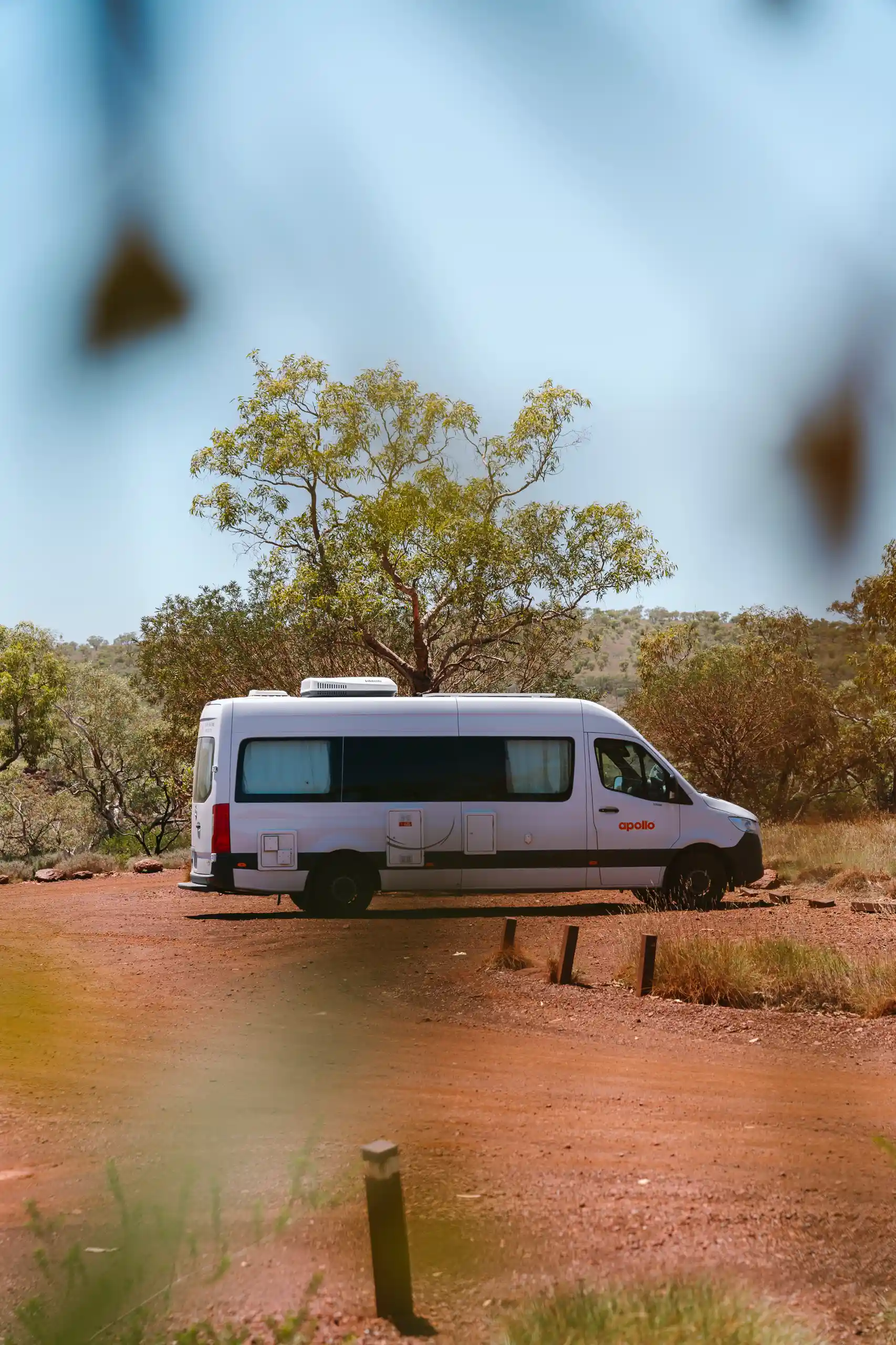 Ein Camper von Apollo parkt im australischen Outback. Man sieht Bäume im Hintergrund und roten Sand.