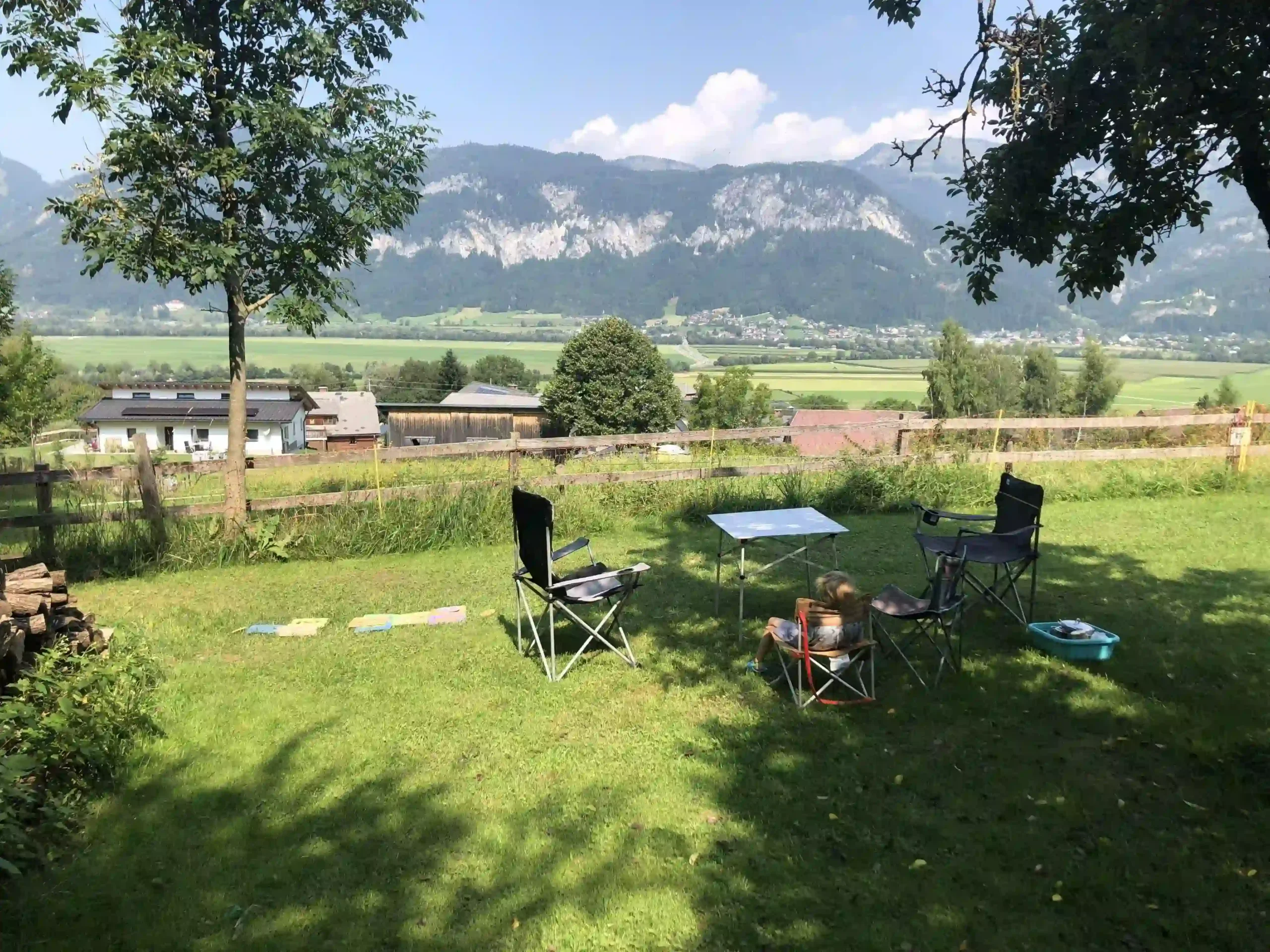 Eine grüne Obstwiese mit einer aufgebauten Camping-Sitzgruppe (Tisch und Stühle). Im Hintergrund ein herrlicher Weitblick über ein grünes Tal.
