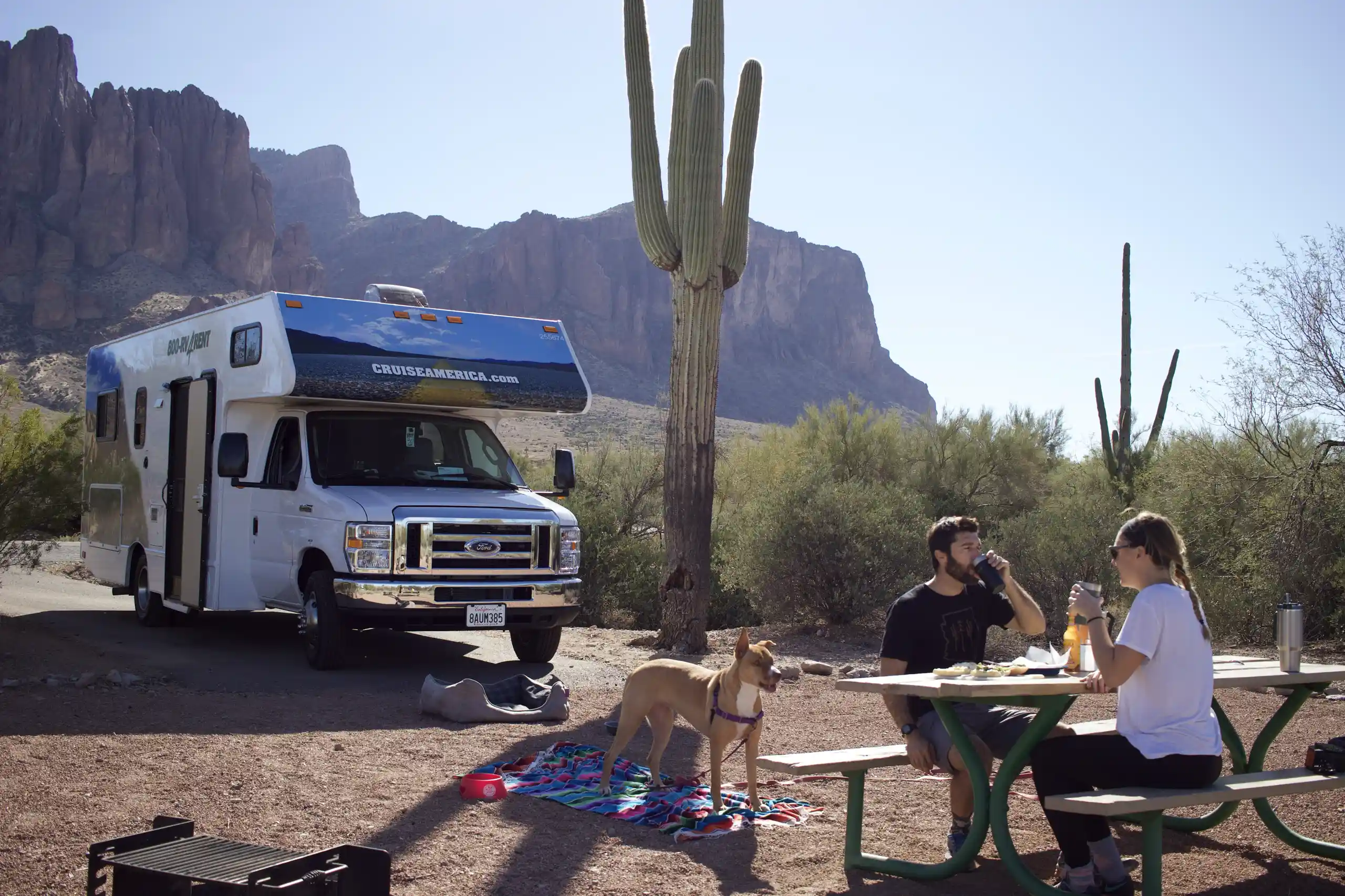Ein Paar sitzt am Tisch auf dem Campingplatz in den USA mit ihrem Hund auf einer Decke vor einem Wohnmobil von Cruise America.