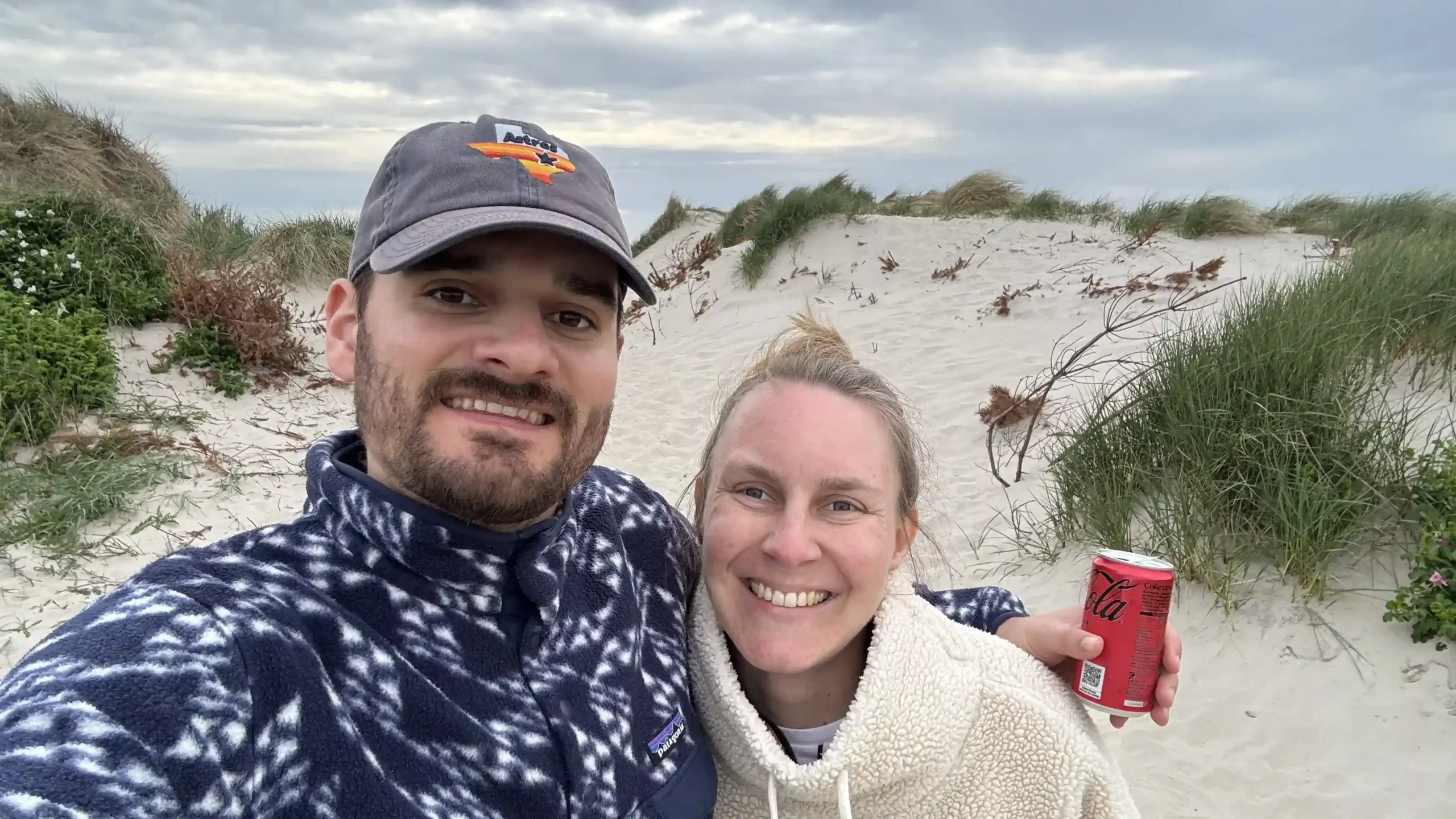 Selfie von einem glücklich lächelnden Paar, das in einer mit Dünengras bewachsenen Sanddüne steht.