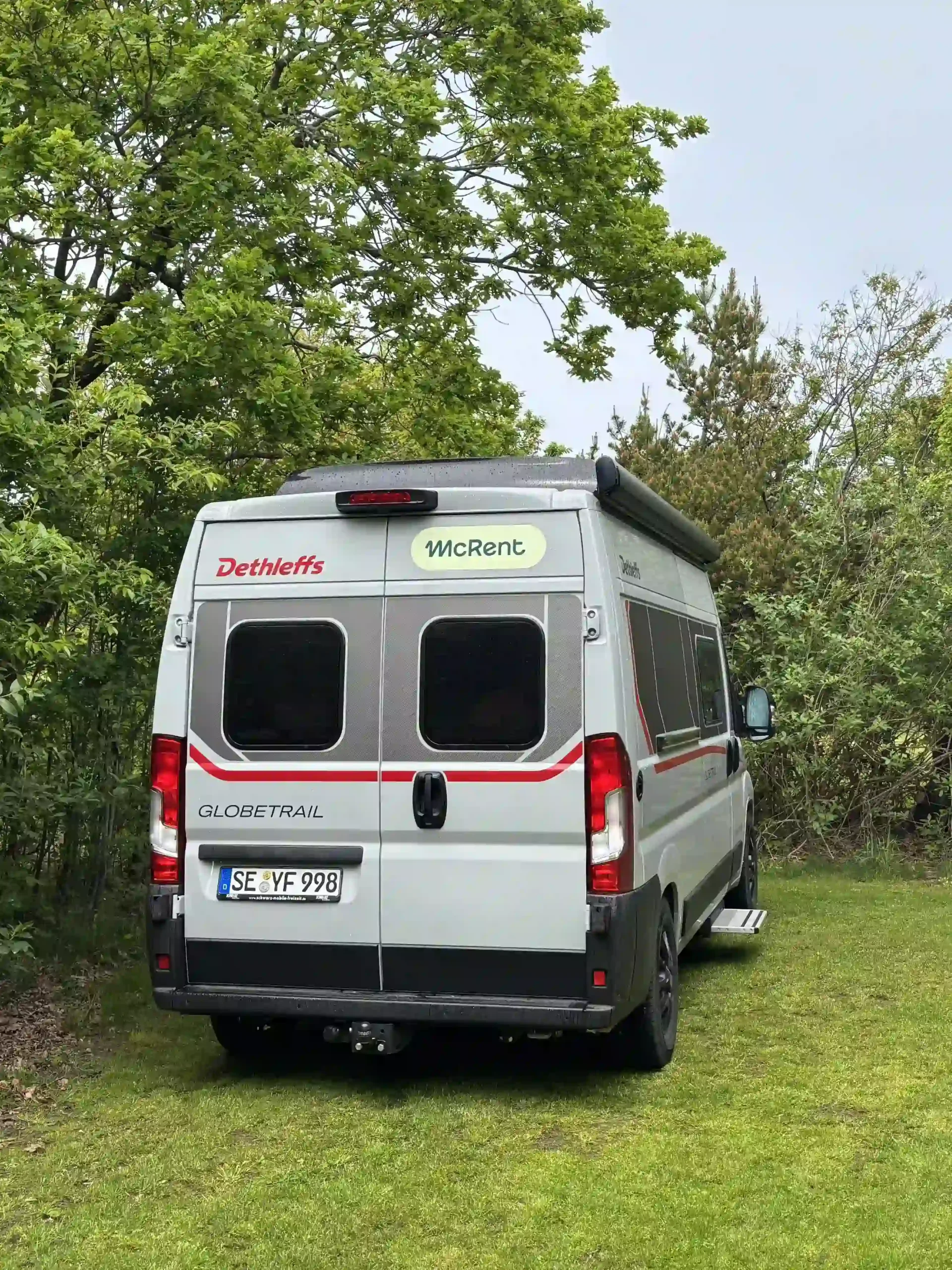 Eine Heckansicht eines silbernen wohnmobils, geparkt auf Rasen vor dichten grünen Bäumen. Die rechte Seite zeigt eine ausgefahrene Trittstufe und eine eingefahrene Markise.