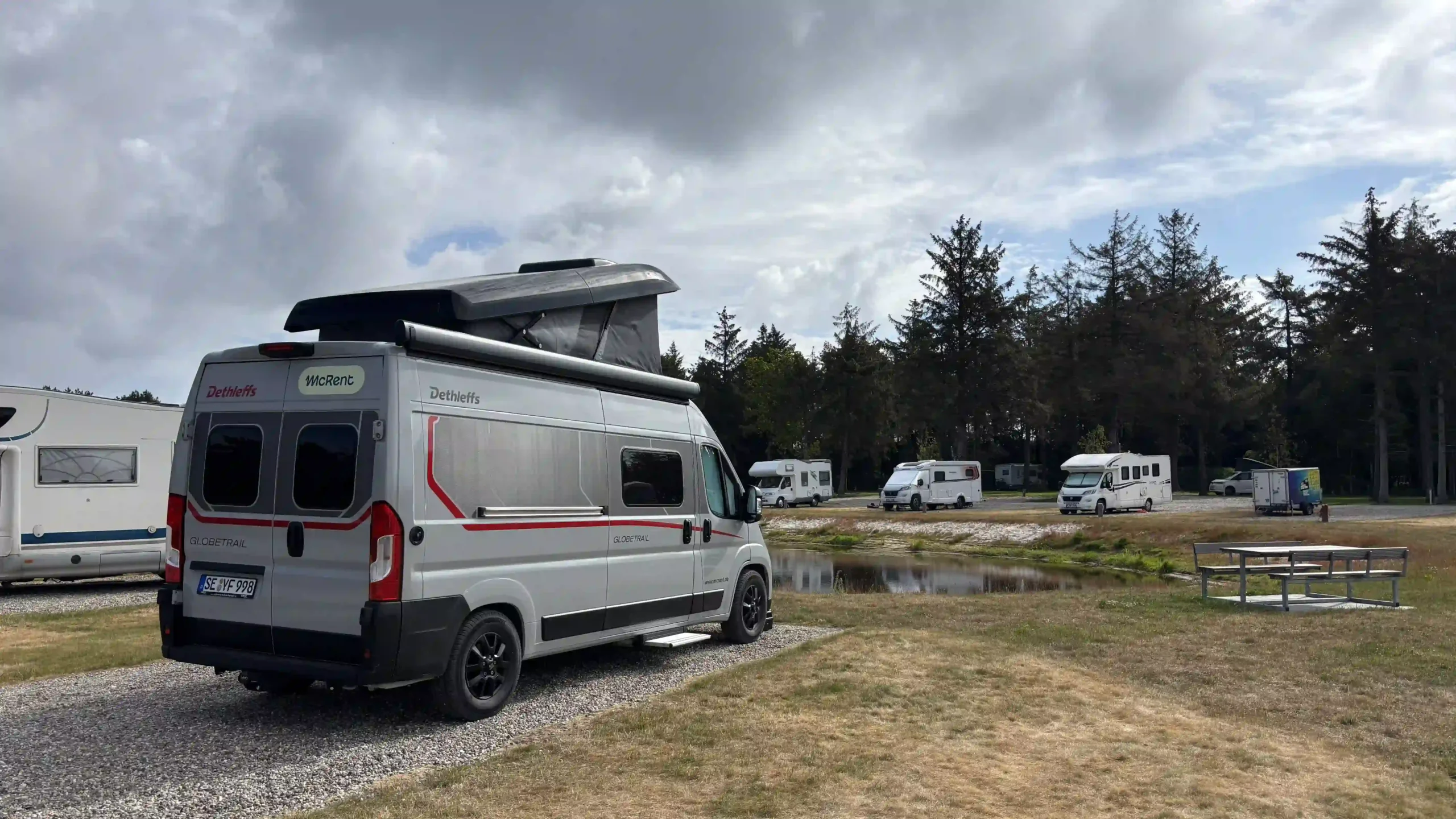 Wohnmobil mit aufgeklapptem Schlafdach steht auf einem Schotterstellplatz. Im Hintergrund befindet sich ein kleiner Teich, weitere geparkte Reisemobile und eine dicht bewachsene Baumliniene.