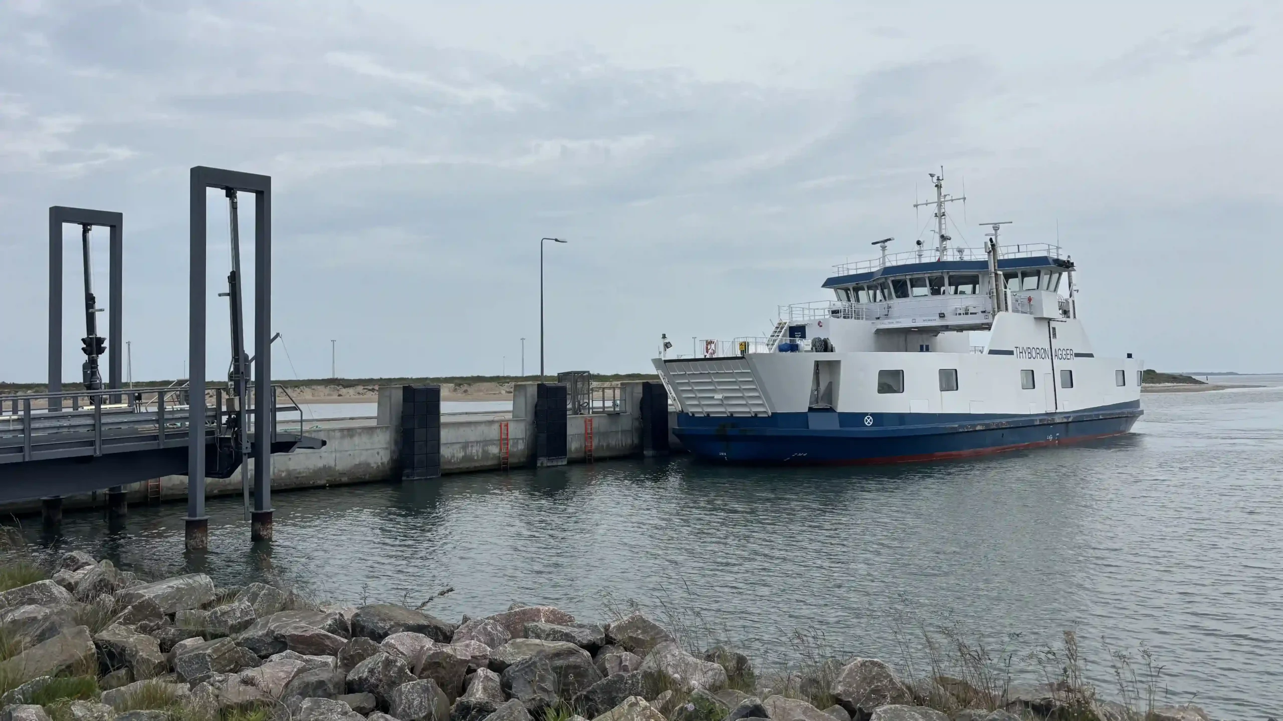 Eine weiß-blaue Autofähre mit der Aufschrift „Thyborøn-Agger“ legt an einem grauen Betonpier an.