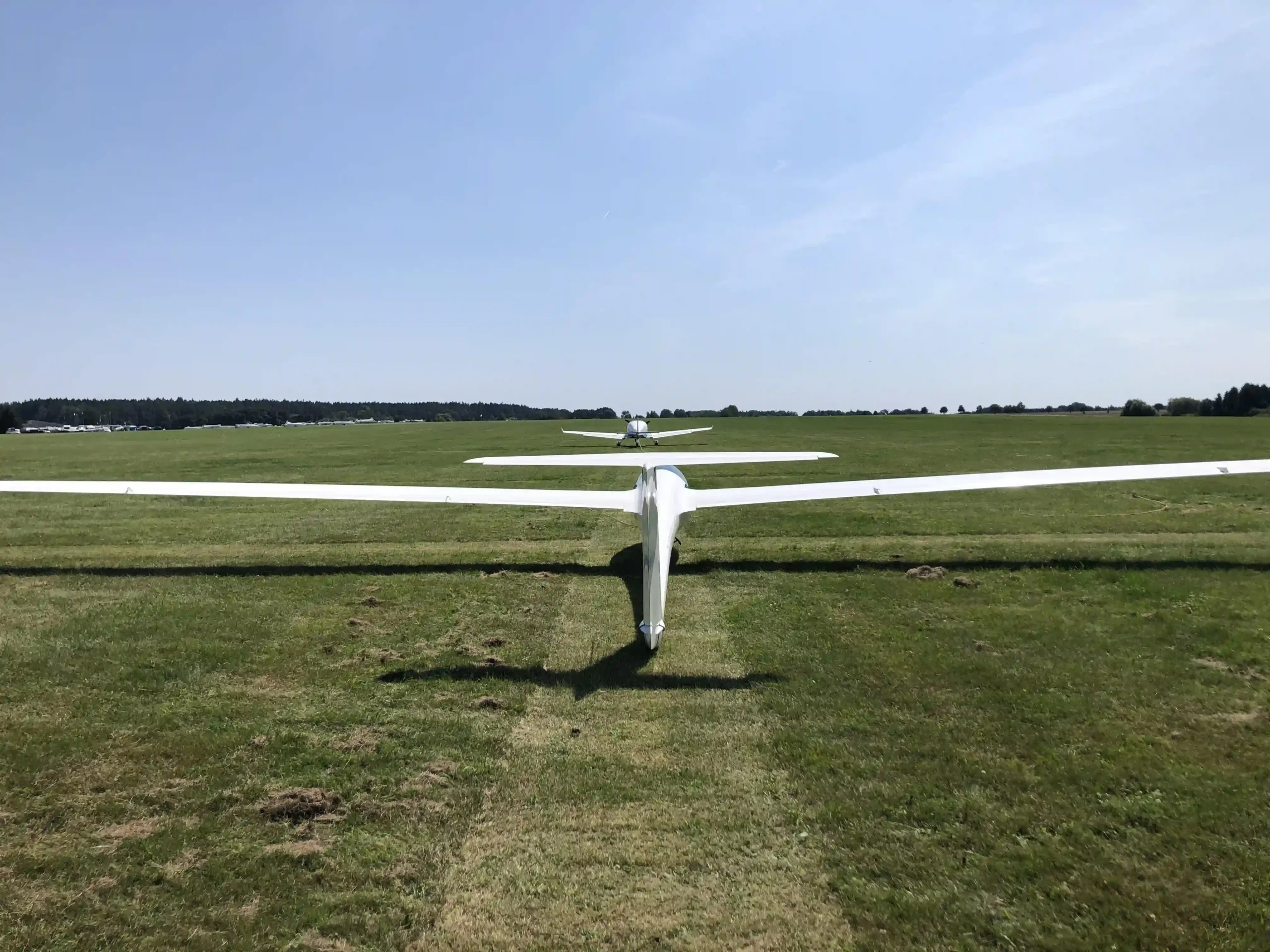 Blick von hinten auf ein weißes Segelflugzeug, das auf einer weiten grünen Grasstartbahn steht.