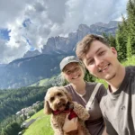 Zwei lächelnde Personen mit einem Hund auf dem Arm, die vor der Kulisse von Gebirgen stehen.