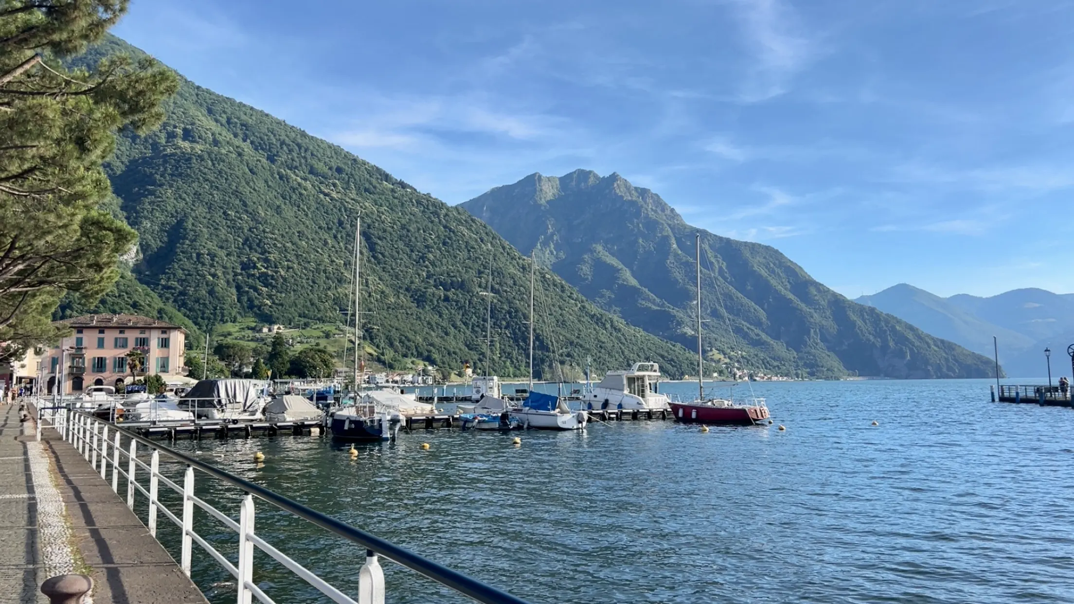 Im Vordergrund zu sehen ist der Bootsanleger des hellblauen Sees Lago de Iseo mit einigen Booten, dahinter Gebirge bei hellblauem Sonnenschein.