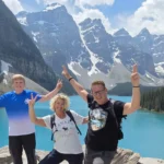 Drei lächelnde, die Arme in die Höhe streckenden Personen posierend vor dem kristallklaren Moraine Lake in Banff. Im Horizont türmen sich die an den Spitzen mit Schnee bedeckten grauen Berge.