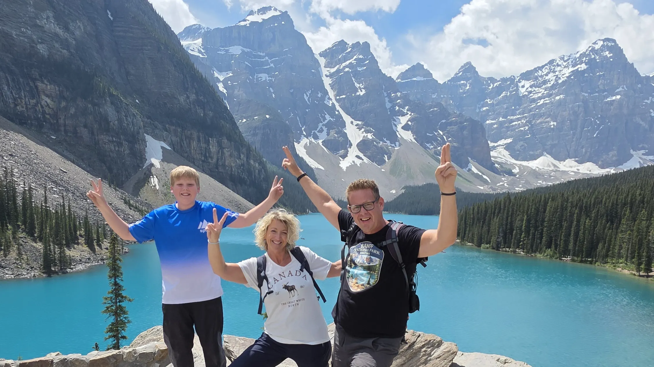 Drei lächelnde, die Arme in die Höhe streckenden Personen posierend vor dem kristallklaren Moraine Lake in Banff. Im Horizont türmen sich die an den Spitzen mit Schnee bedeckten grauen Berge.