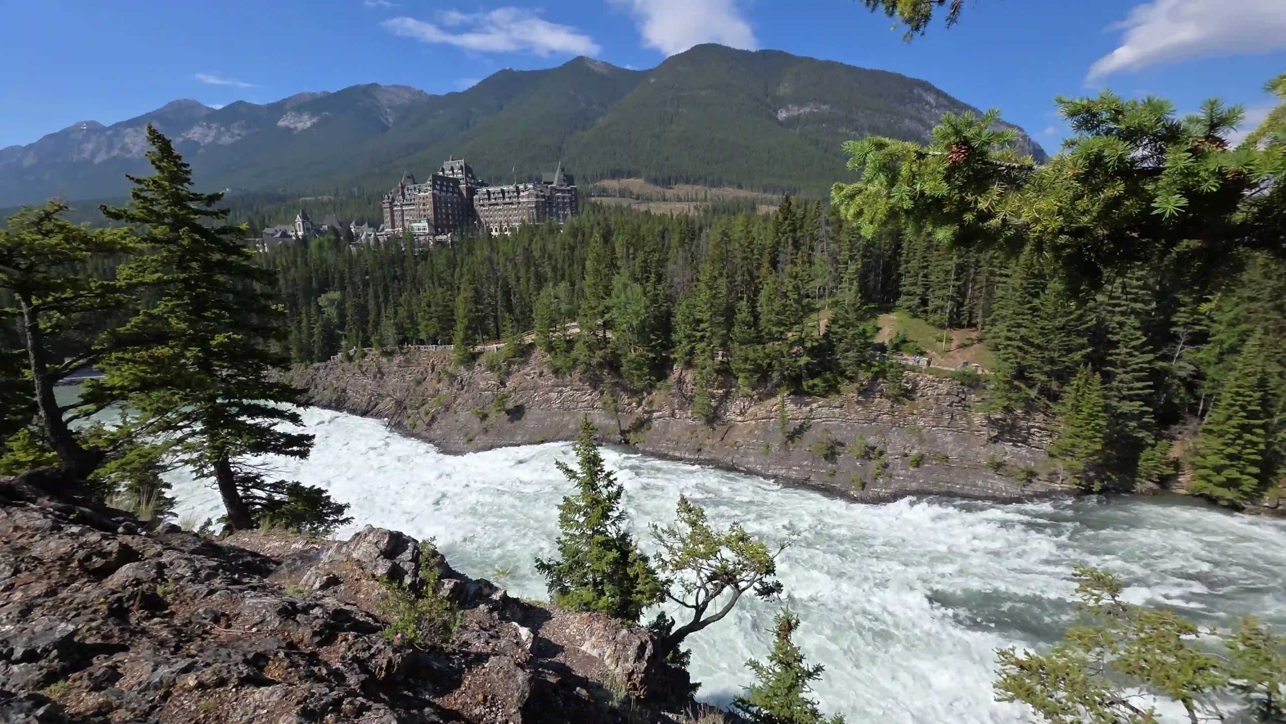 Die Vogelperspektive auf einen durch das Bild fließenden Fluss mit starker Strömung. Der Fluss ist umgeben von Tannen und Gestein. Im Hintergrund ragt das Banff Springs Hotel aus dem Grün der Tannen.