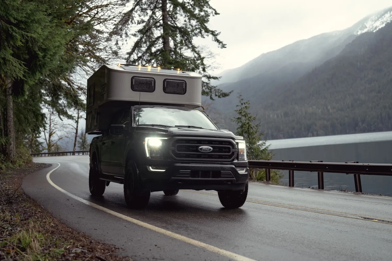 Ein dunkler Truck Camper fährt auf einer kurvigen Straße entlang eines nebligen Bergsees in Kanada.
