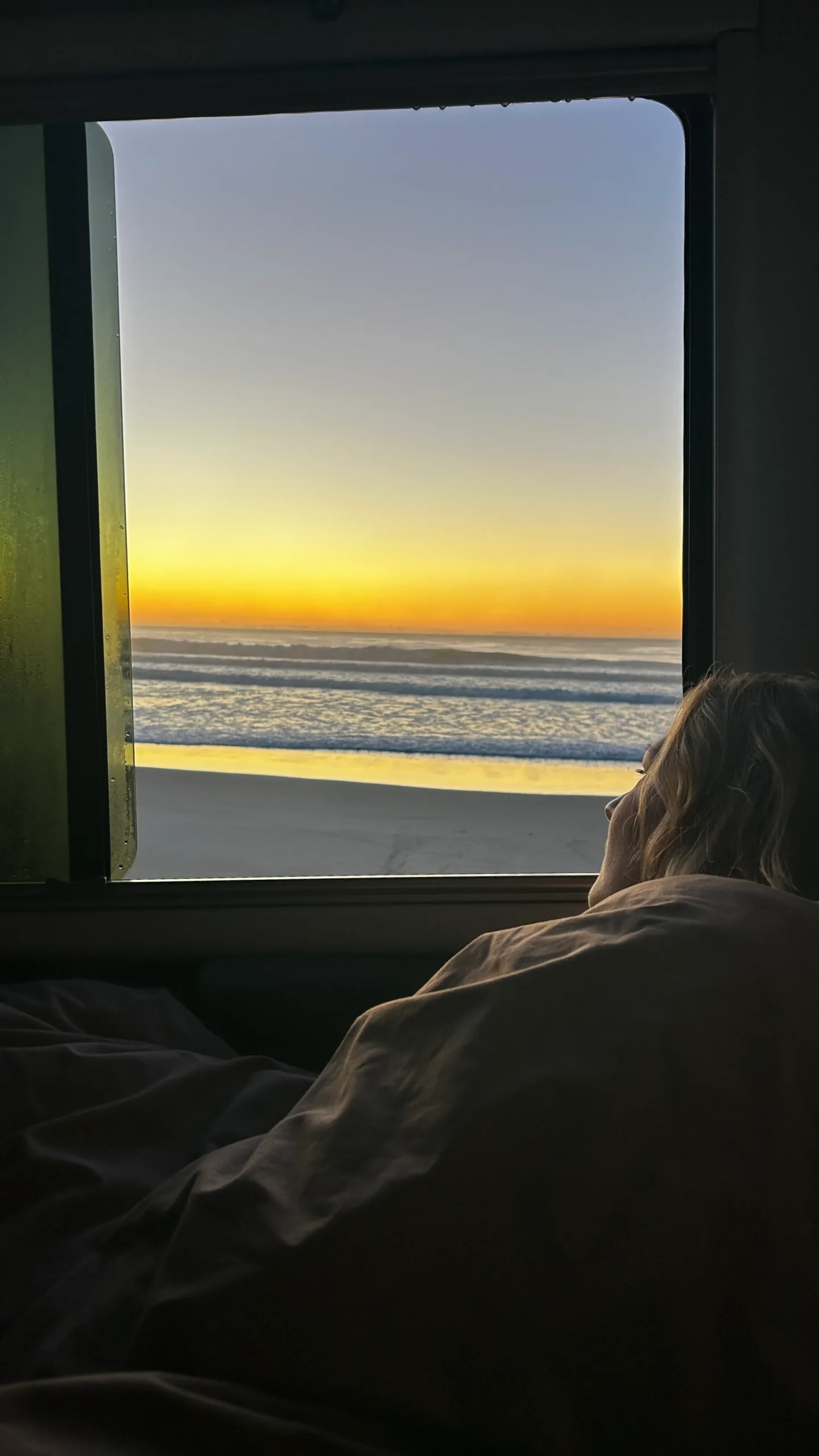 Blick aus einem Wohnmobilfenster auf einen goldenen Sonnenaufgang über dem Meer. Im Vordergrund liegt eine Frau gemütlich eingekuschelt in Decken und schaut auf die Wellen.