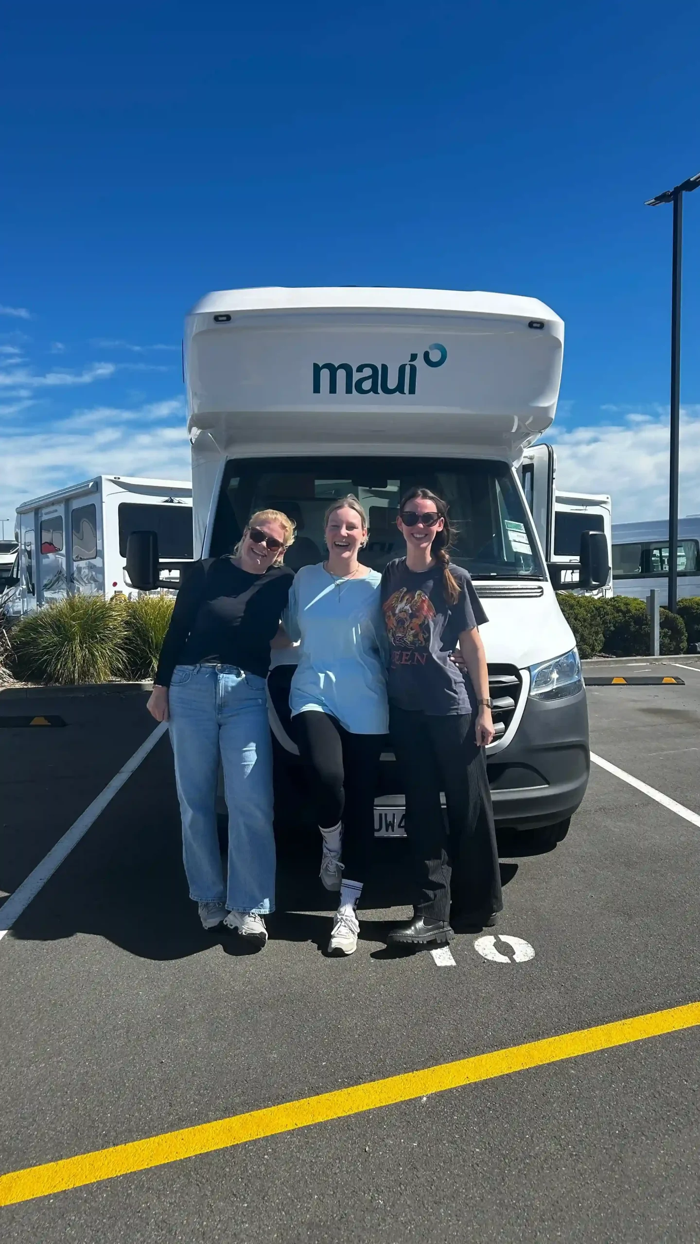 Drei strahlende junge Frauen stehen lachend vor einem großen weißen Maui-Camper auf einem Parkplatz in Christchurch.