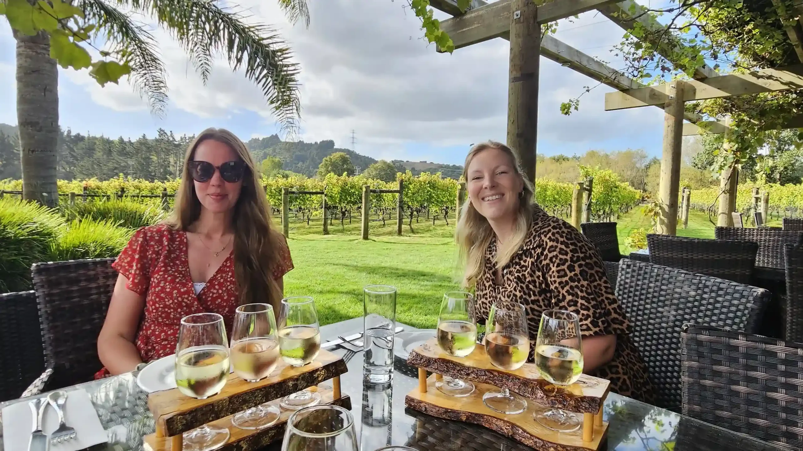 Zwei lächelnde Frauen sitzen an einem Gartentisch in einem Weinberg und genießen ein Wein-Tasting.