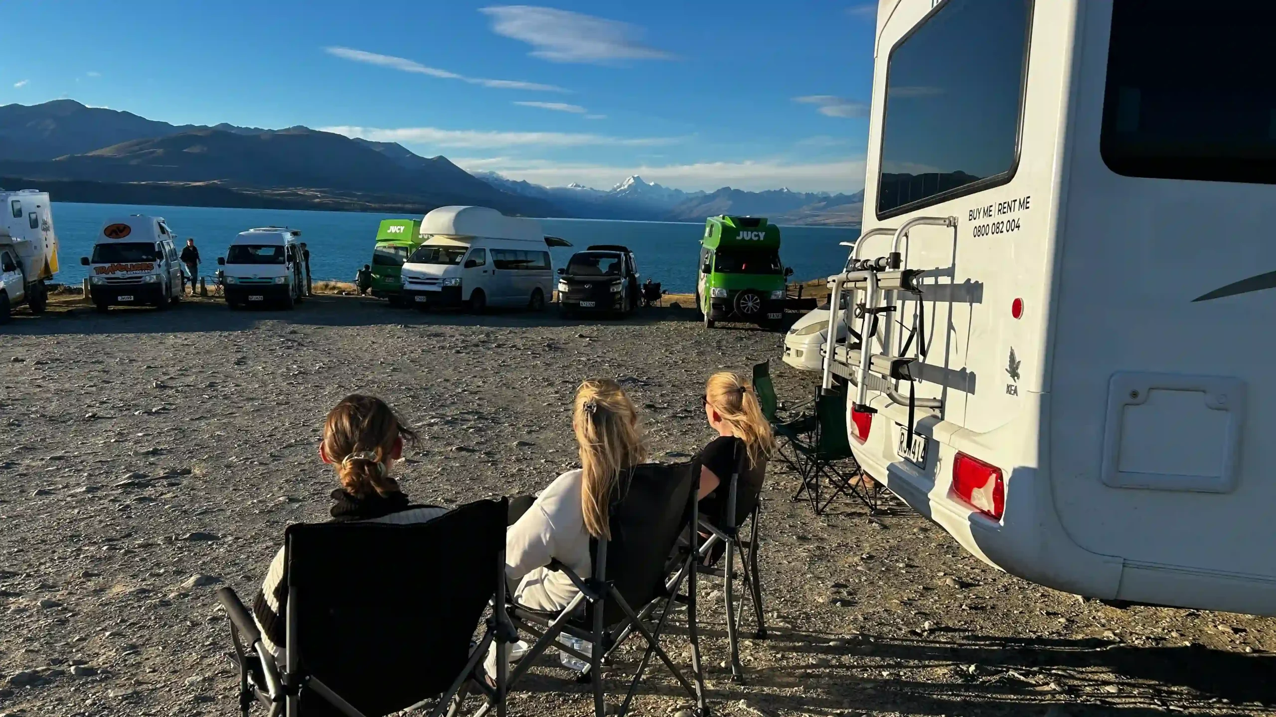 Drei Frauen sitzen in Campingstühlen vor ihrem Wohnmobil und blicken auf den hellblauen Lake Pukaki und die schneebedeckten Südalpen Neuseelands.