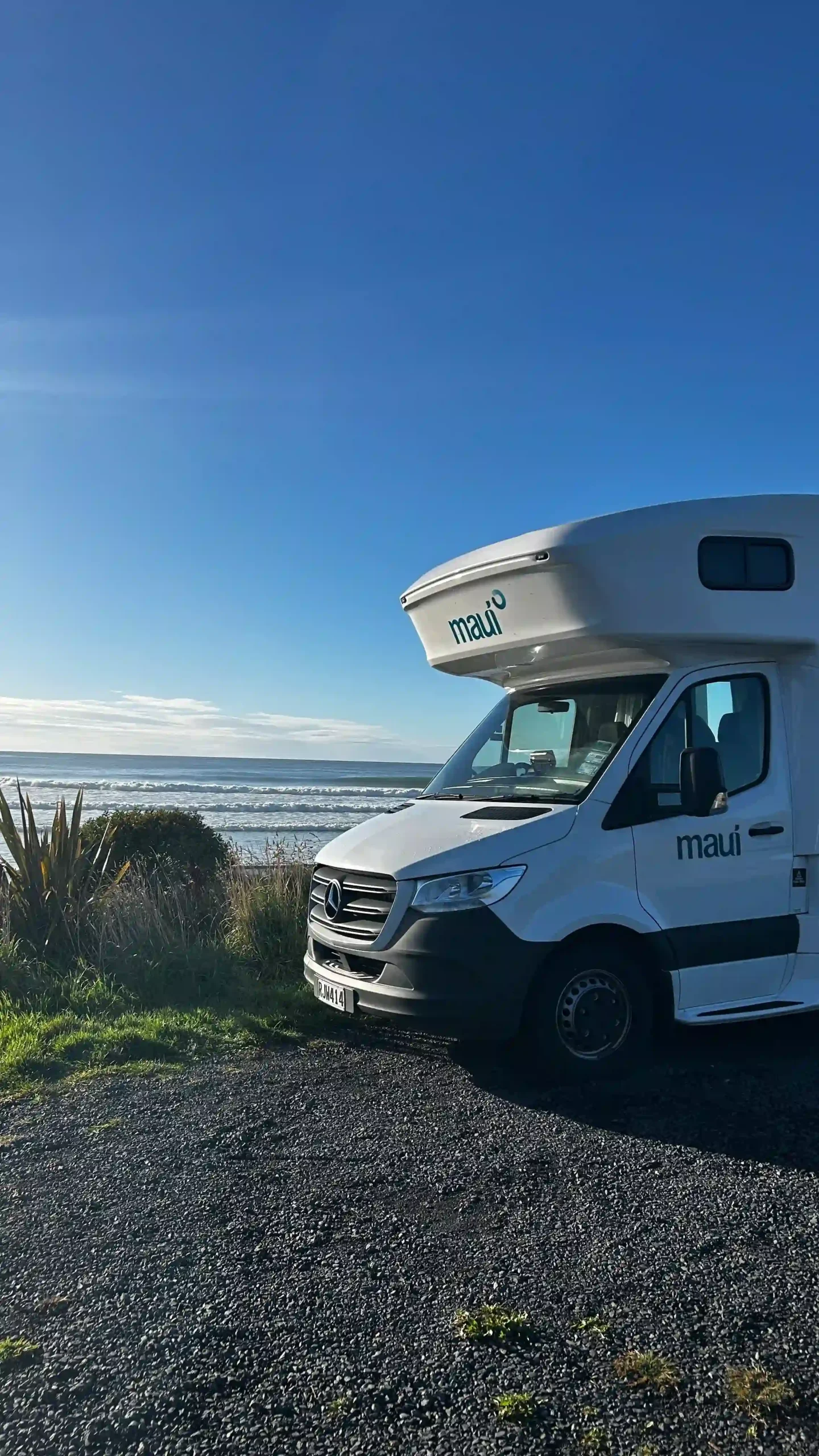 Ein weißer Maui-Camper steht auf einem Schotterplatz direkt vor dem weiten Ozean unter strahlend blauem Himmel.