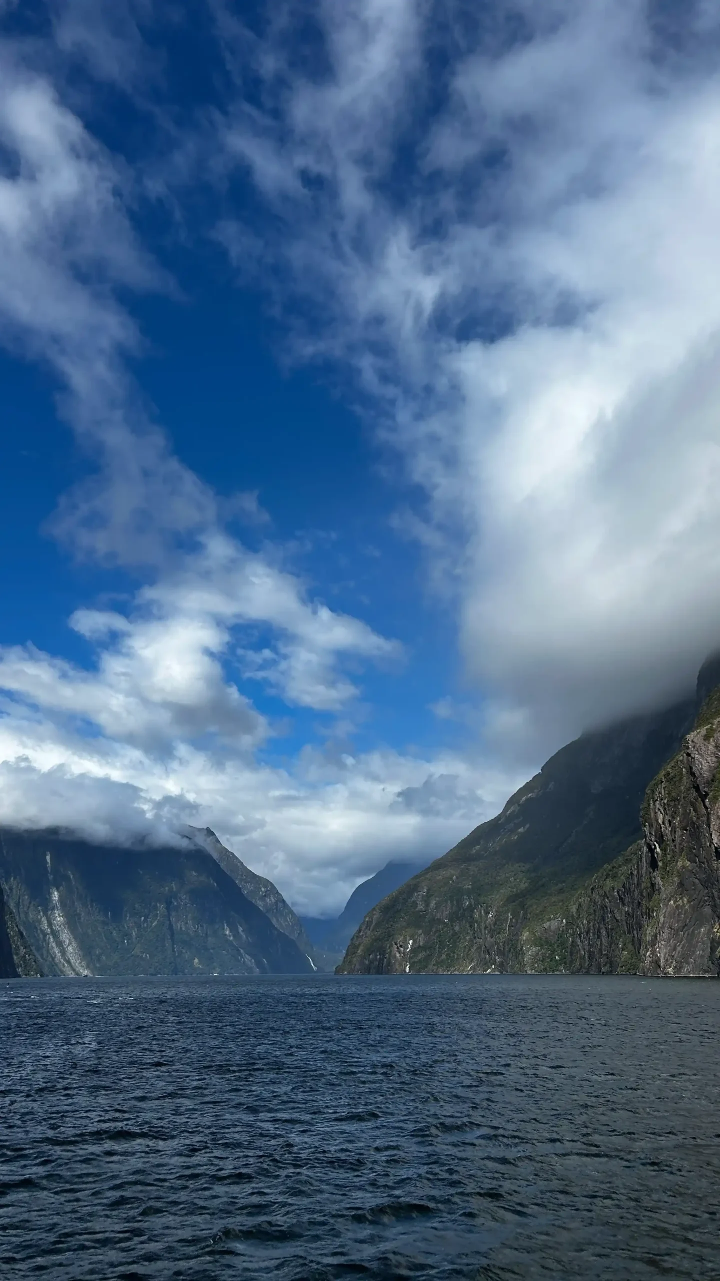 Weite Ansicht des Milford Sound in Neuseeland mit dunklem Wasser, steilen grünen Klippen und tief hängenden weißen Wolken unter blauem Himmel.