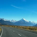 Eine asphaltierte Landstraße führt auf die schneebedeckten Südalpen Neuseelands zu, dominiert vom markanten Gipfel des Mount Cook.