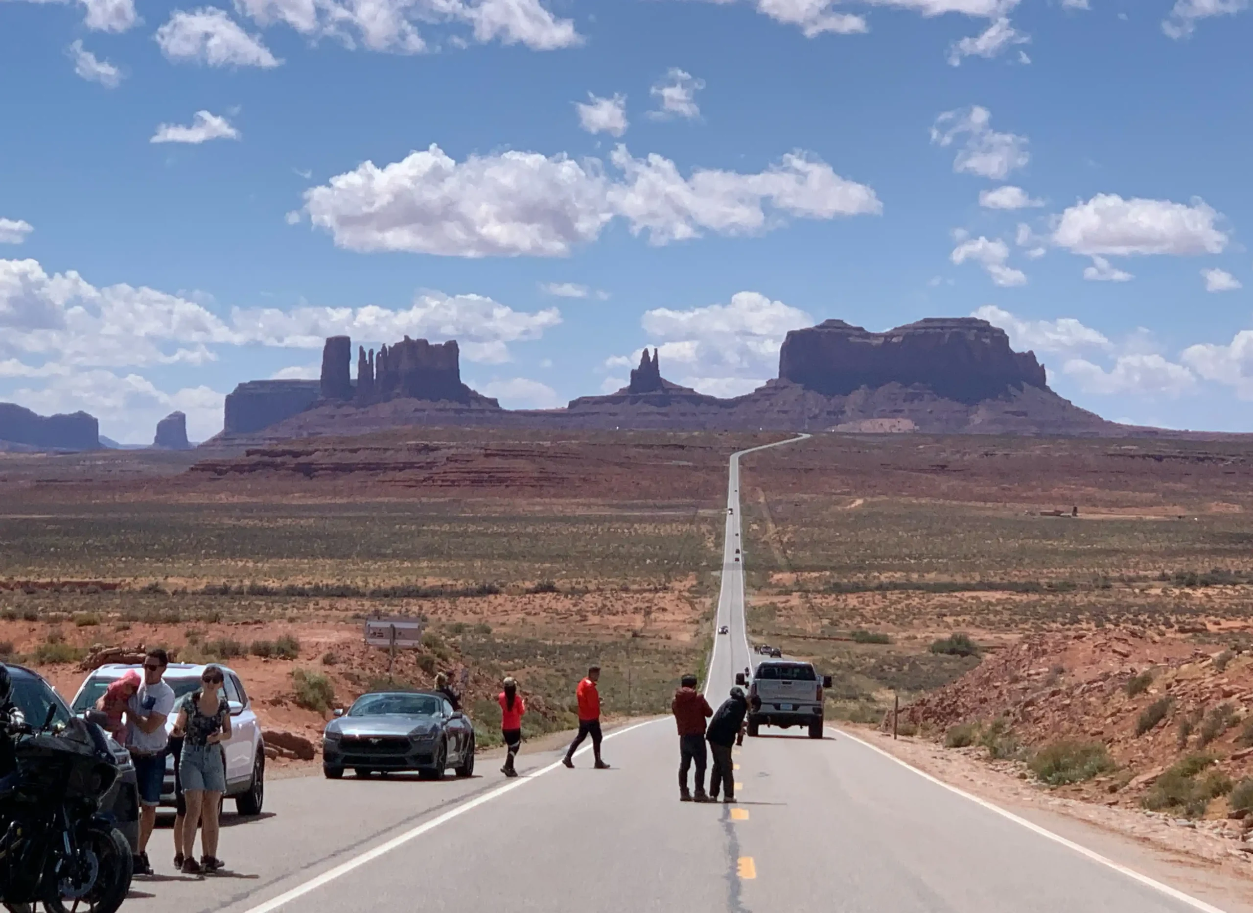 Eine lange, gerade Landstraße führt durch eine rötliche Wüstenlandschaft direkt auf die markanten Berge des Monument Valley zu. Im Vordergrund halten mehrere Touristen am Straßenrand und auf der Fahrbahn, um Fotos von der berühmten Aussicht zu machen.