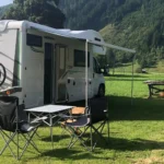 Wohnmobil auf einer grünen Wiese. Im Vordergrund stehen ein Campingtisch und zwei Stühle.