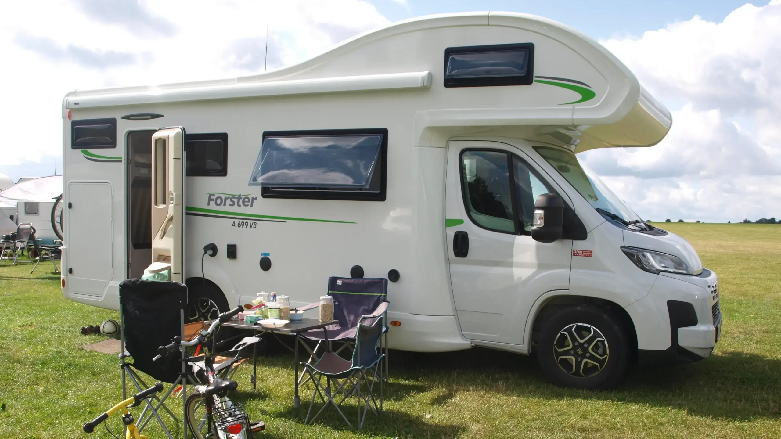 Ein weißes Wohnmobil steht auf einer grünen Wiese unter leicht bewölktem Himmel. Davor ist eine gemütliche Sitzecke mit Campingtisch, Stühlen und Frühstücksutensilien aufgebaut.