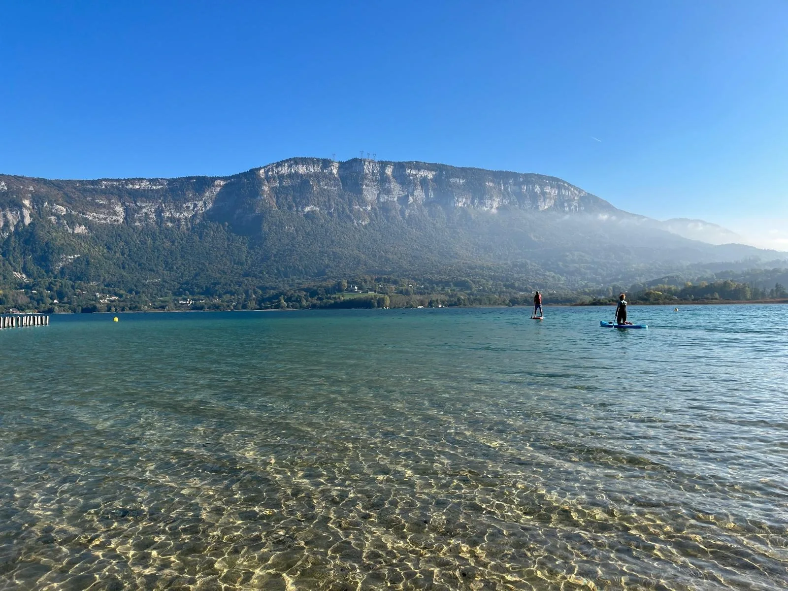 Crystal-clear waters reveal the sandy ripple patterns below as paddleboarders head toward a misty mountain landscape.