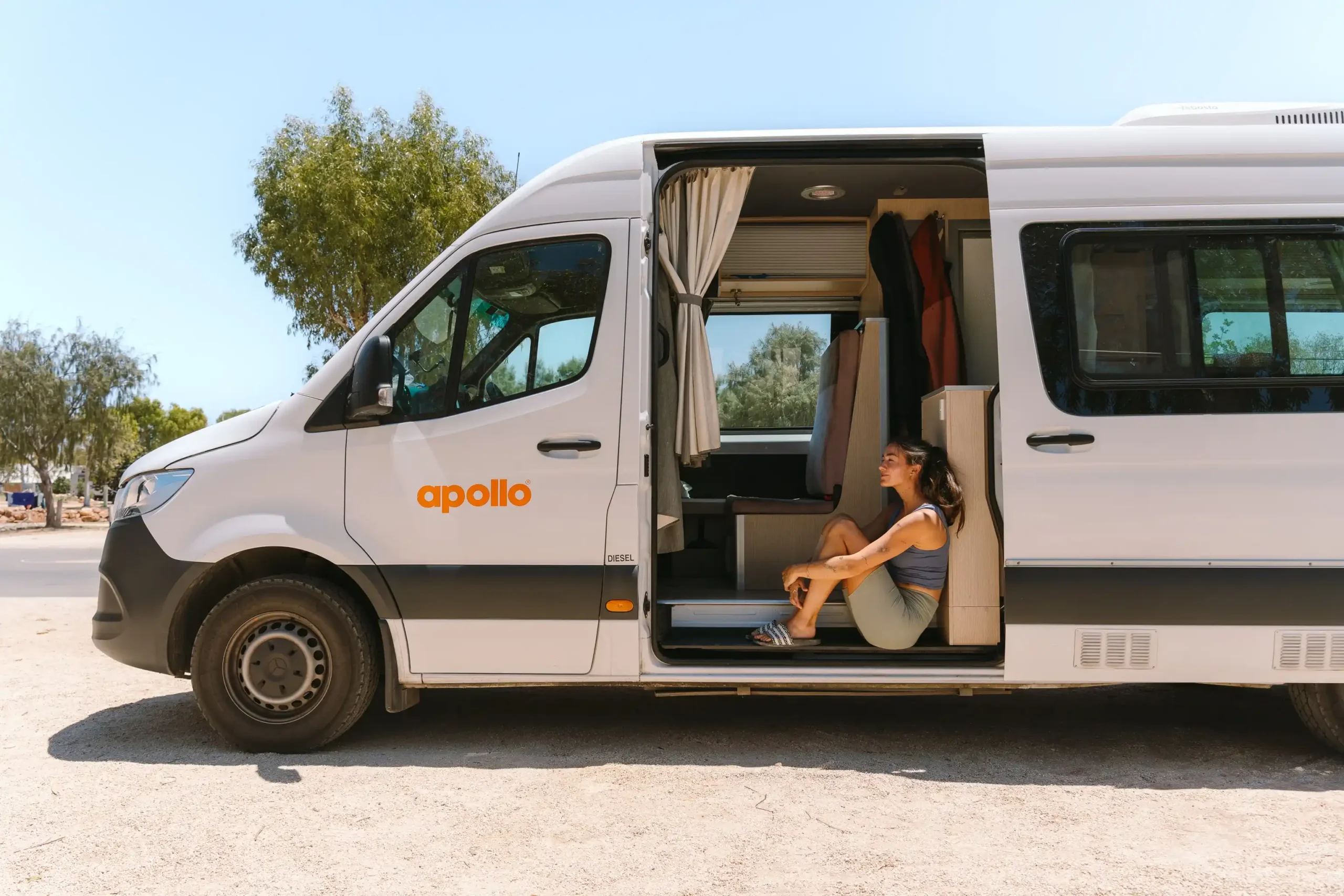 A woman sitting in the open side door of a parked up white Apollo campervan.