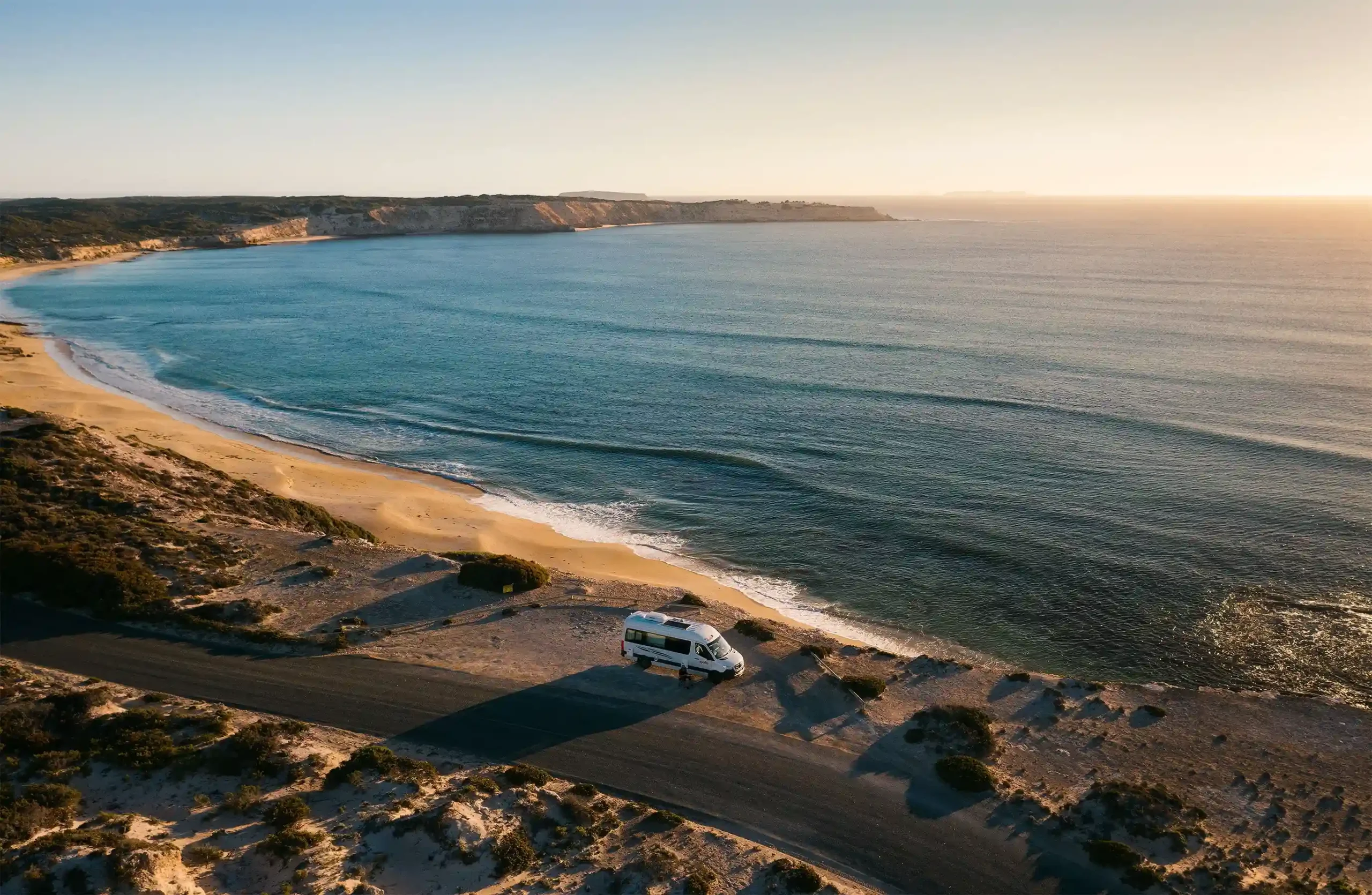 A white campervan is parked on a narrow strip of land between a coastal road and a sandy beach.