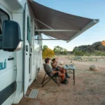 A couple sits in camping chairs under the awning of a white Maui motorhome, enjoying drinks at a small table.