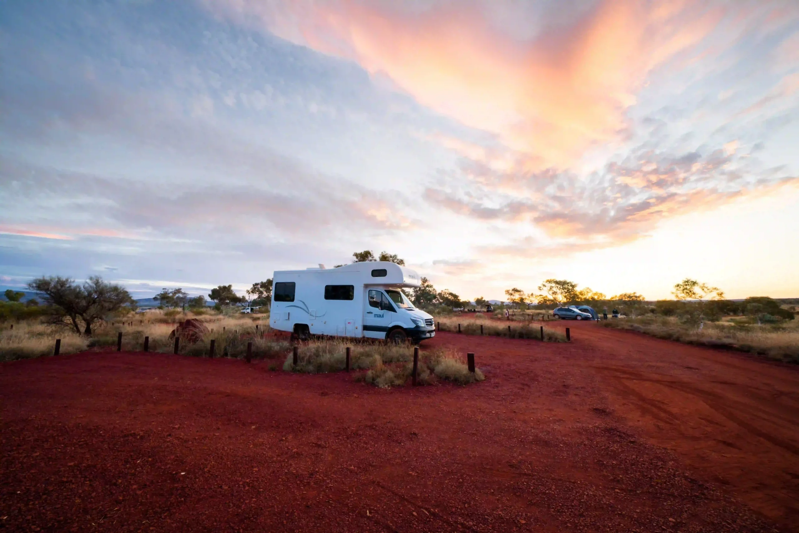 A white Maui motorhome parked up at a red-sand campsite in the Australian outback during a vibrant pink and orange sunset.