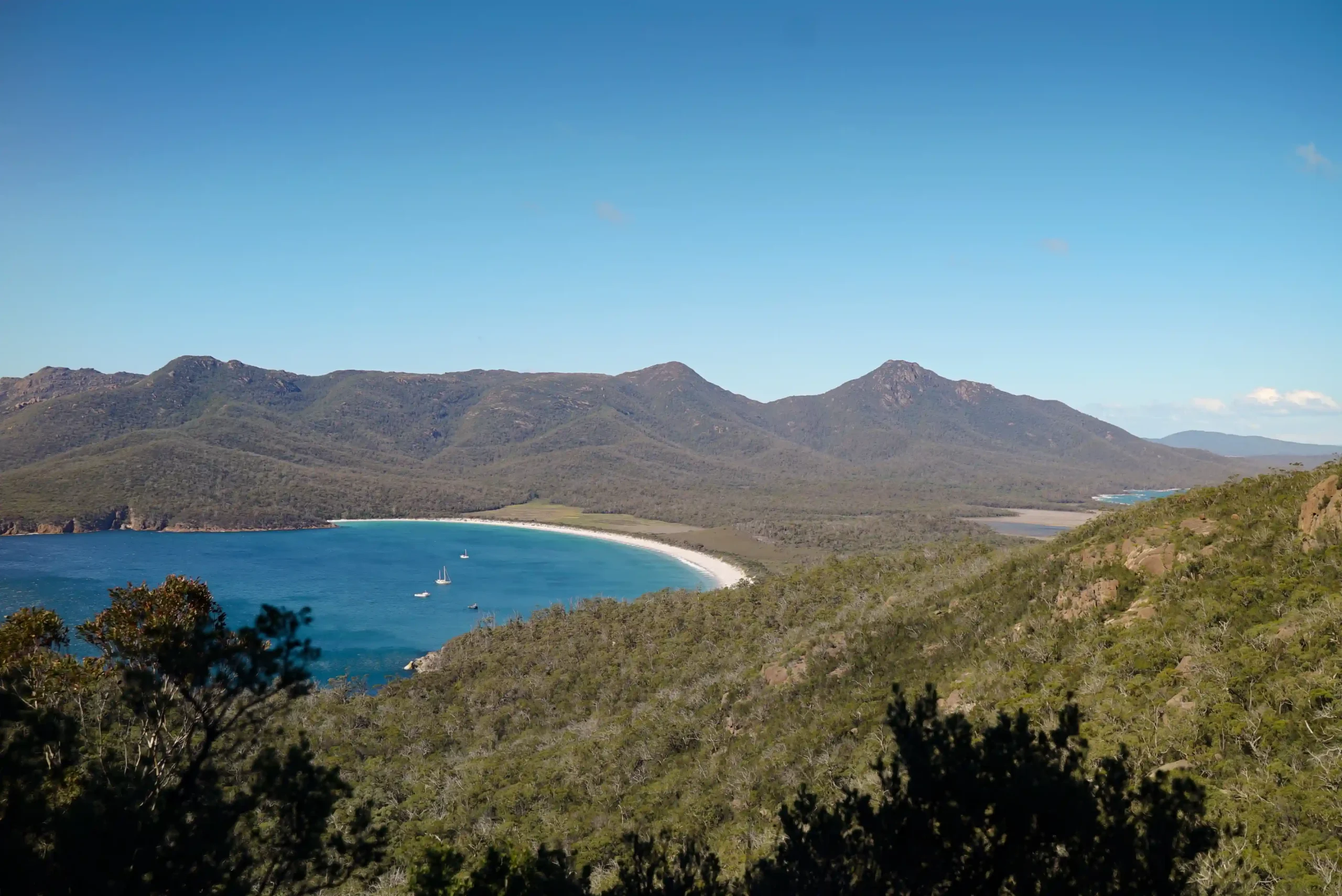 A white sandy beach curves around a bright blue bay, surrounded by dense green forest and rugged mountains under a clear blue sky.