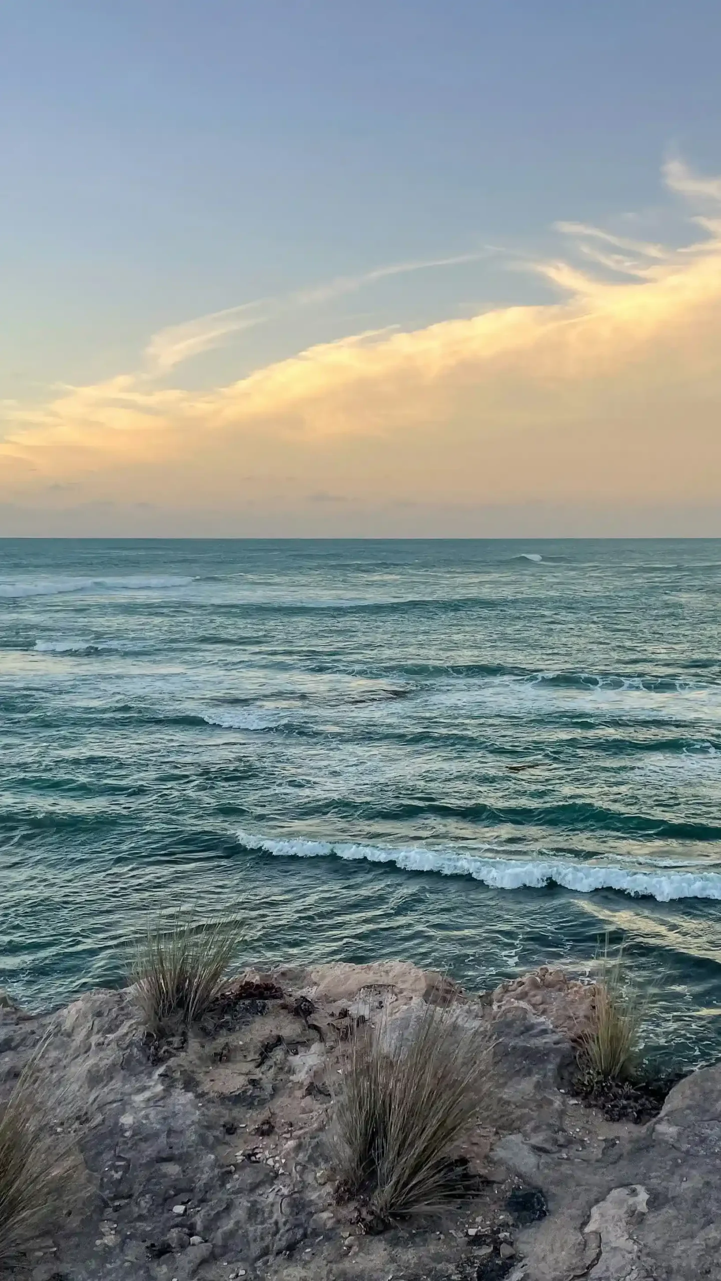 A rocky limestone cliff overlooking a turquoise ocean with white-capped waves. The sky above is a soft gradient of blue and pale orange with wispy, golden-lit clouds during sunset.