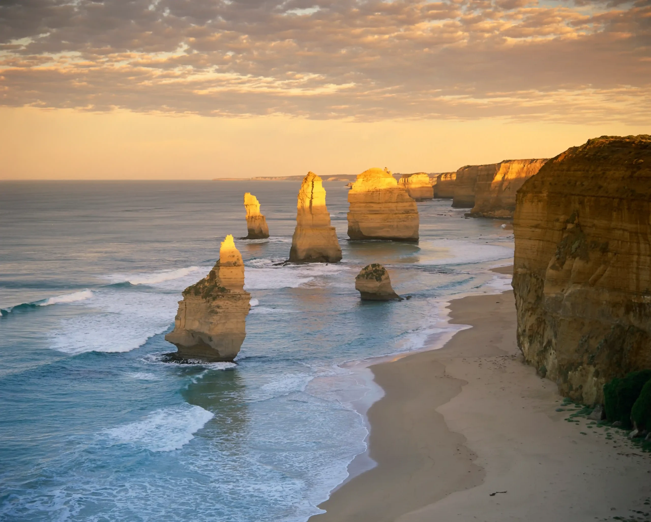 The tall stone pillars rise from the turquoise ocean beside a sandy beach and steep cliffs.