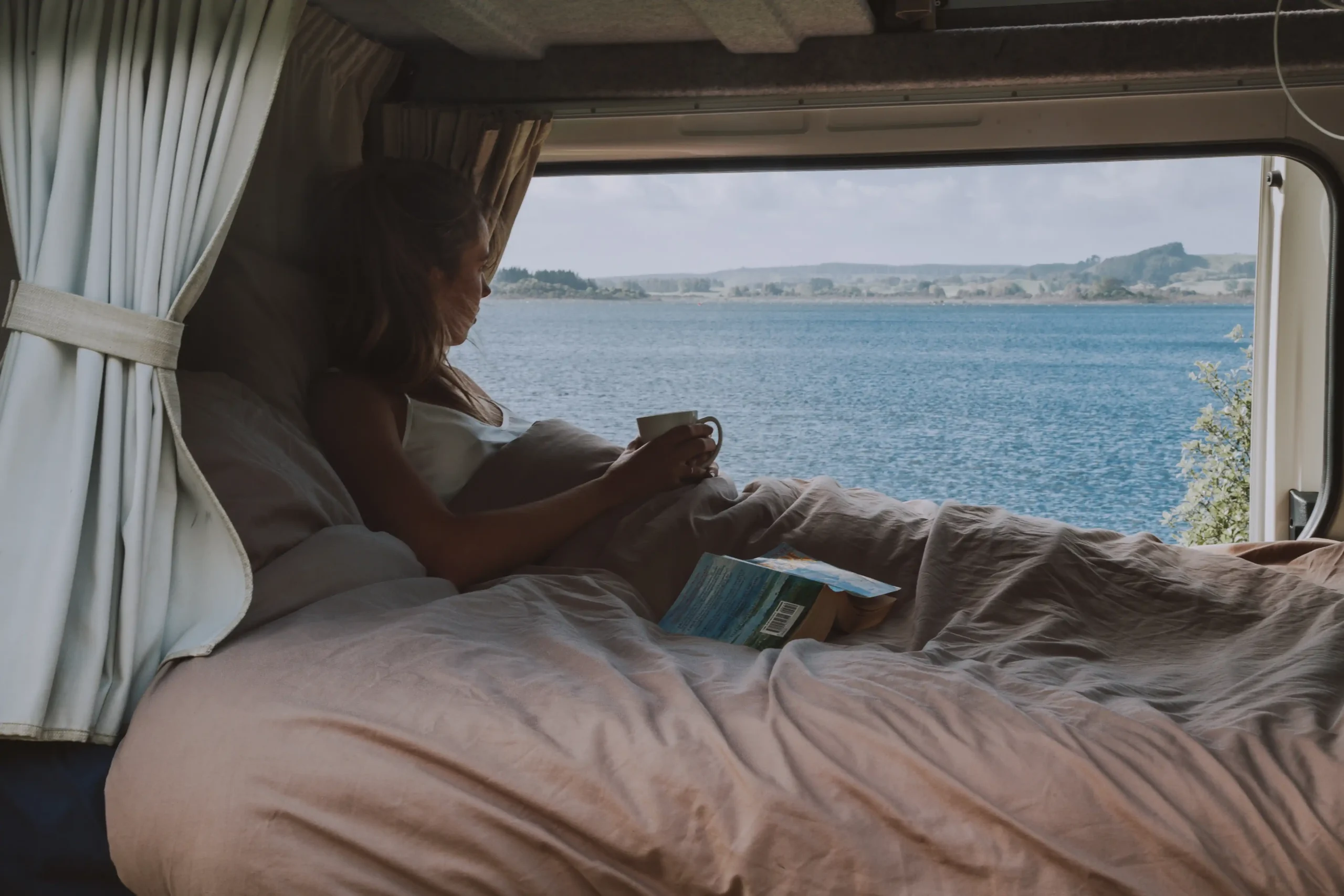 View of a cozy campervan where a woman lies in bed, holding a mug and looking out at the ocean.