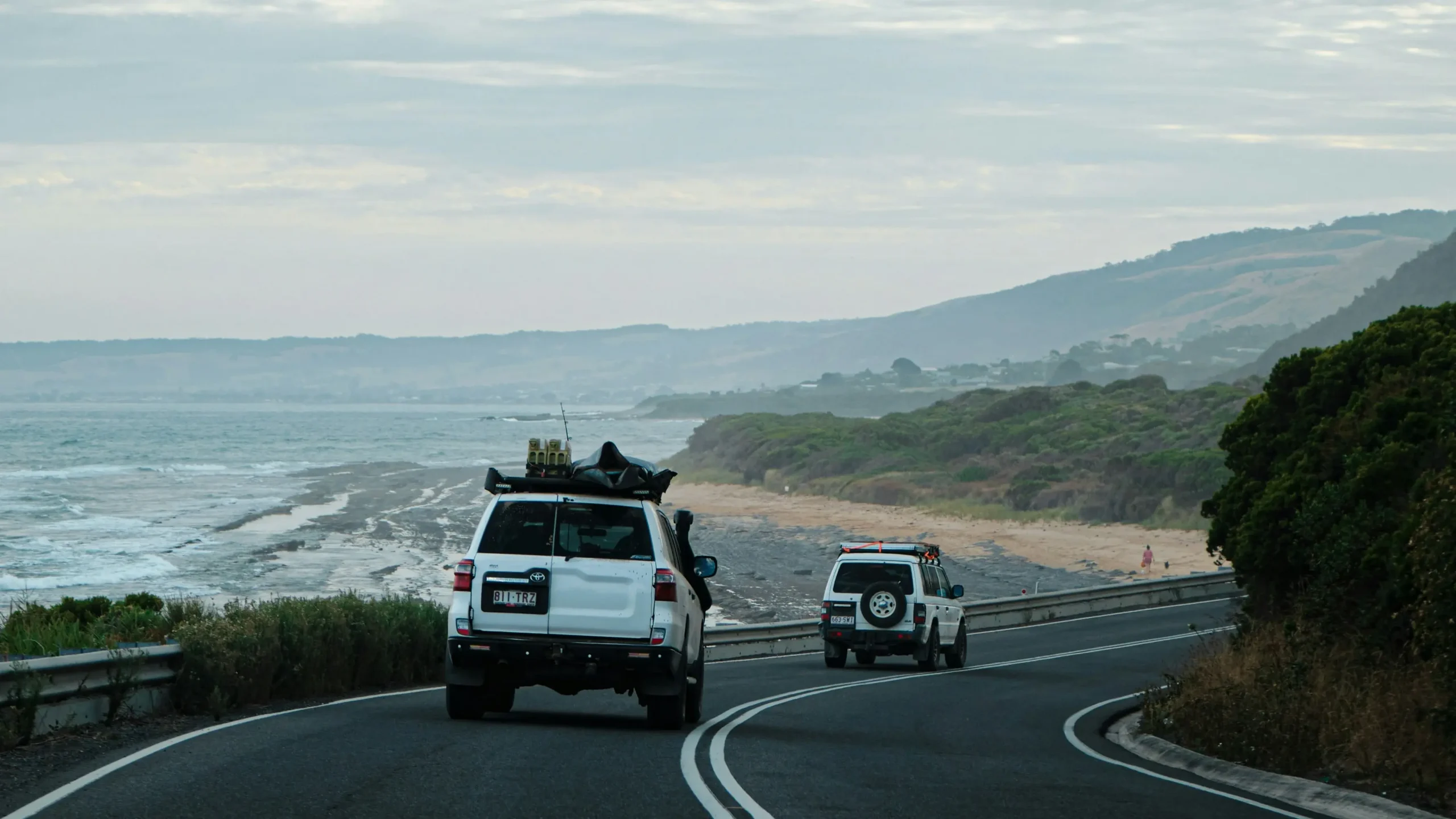 Two white 4WD vehicles equipped with roof racks and camping gear drive along a winding coastal highway.