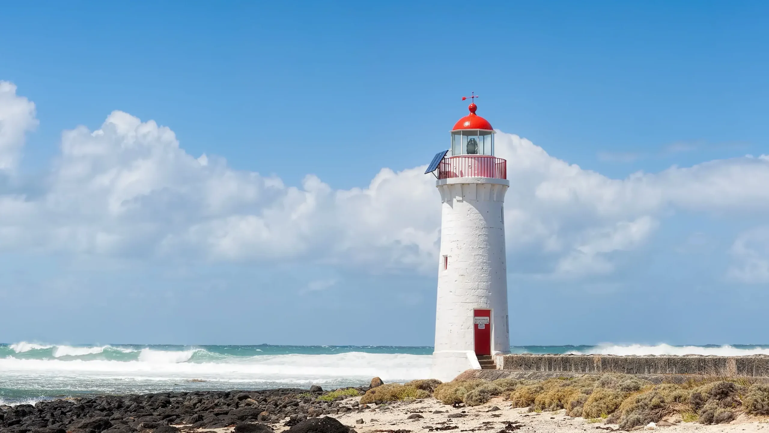 A tall, white stone lighthouse with a vibrant red lantern room and matching red door stands on the rocky coast of Port Fairy, Victoria. Crashing white waves of the turquoise ocean meet a dark basalt shoreline under a bright blue sky with fluffy white clouds.