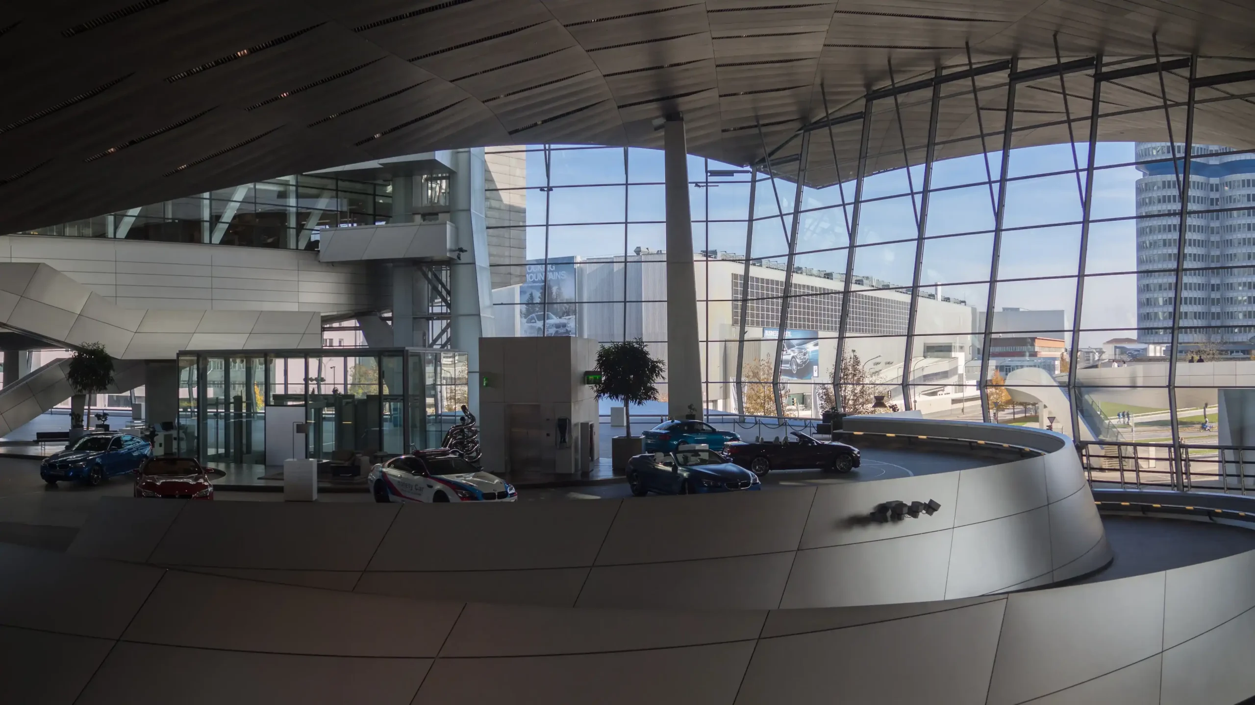 Interior view of the futuristic BMW Welt building in Munich, featuring a curved ramp with luxury cars on display and the BMW headquarters visible through a large glass window.