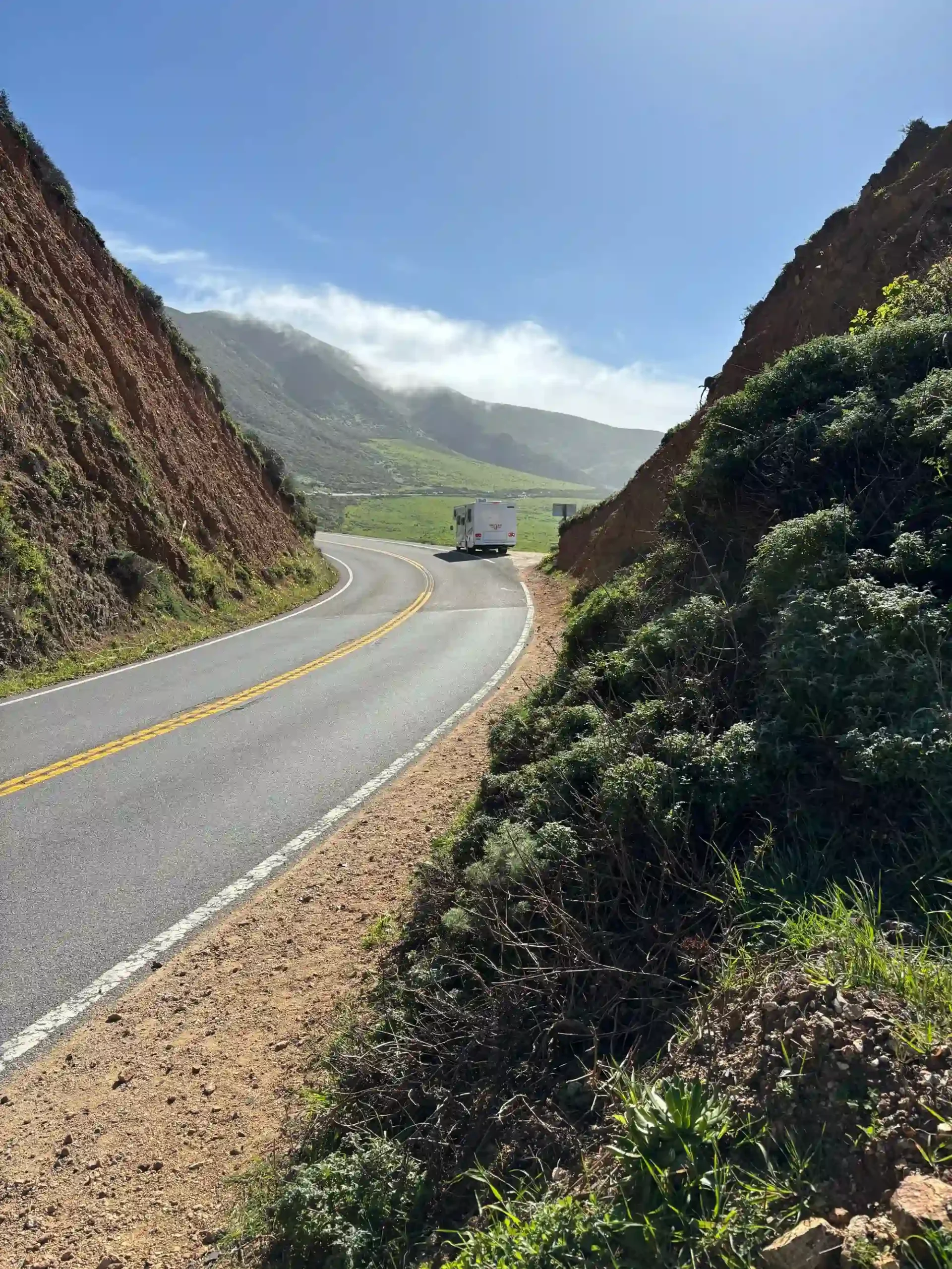 A white motorhome driving around a bend on a highway between steep dirt cliffs, with green rolling hills and misty mountains under a bright sun.