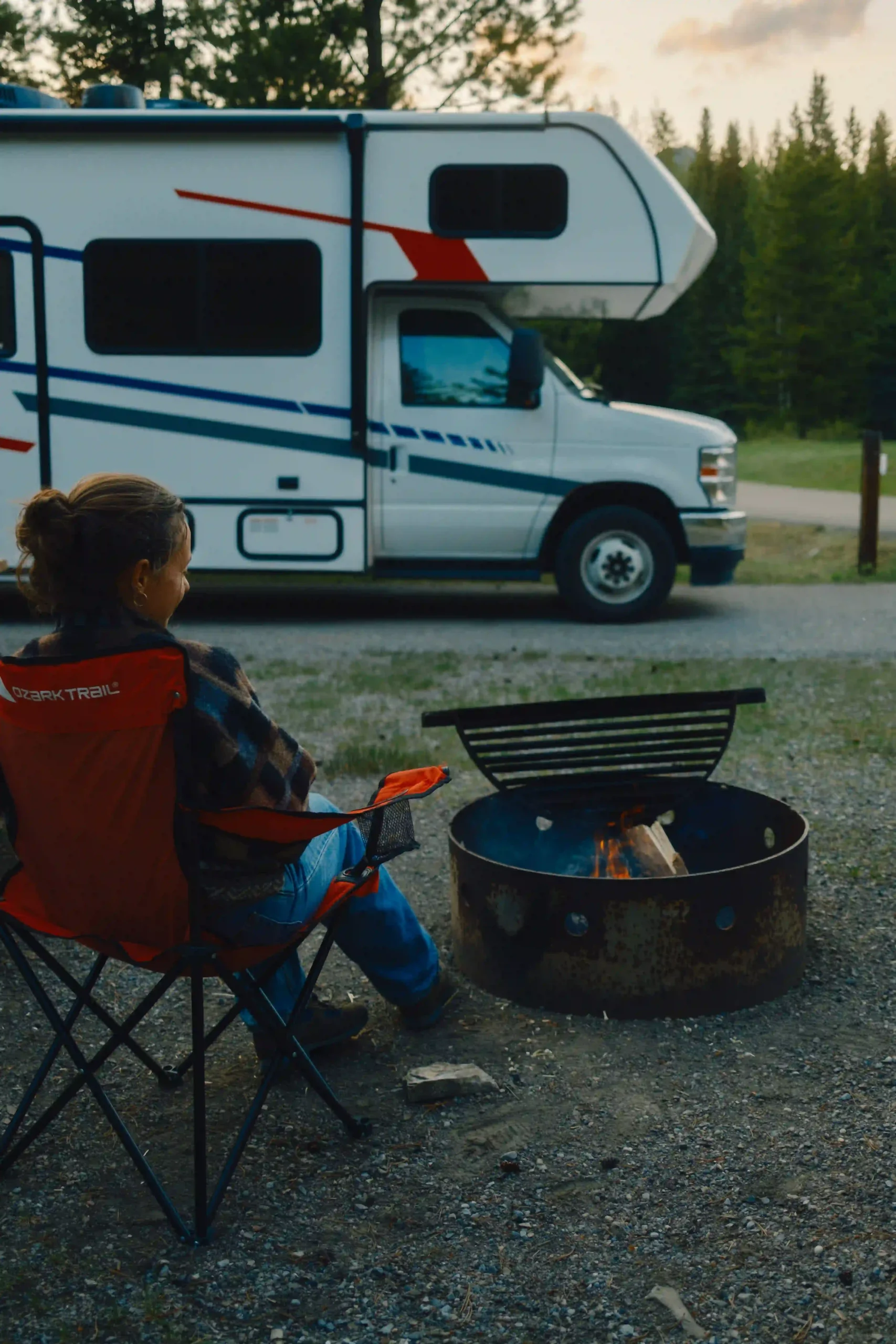 A person sits in a camping chair beside a lit fire pit, facing a parked motorhome at a forest campsite during early evening.