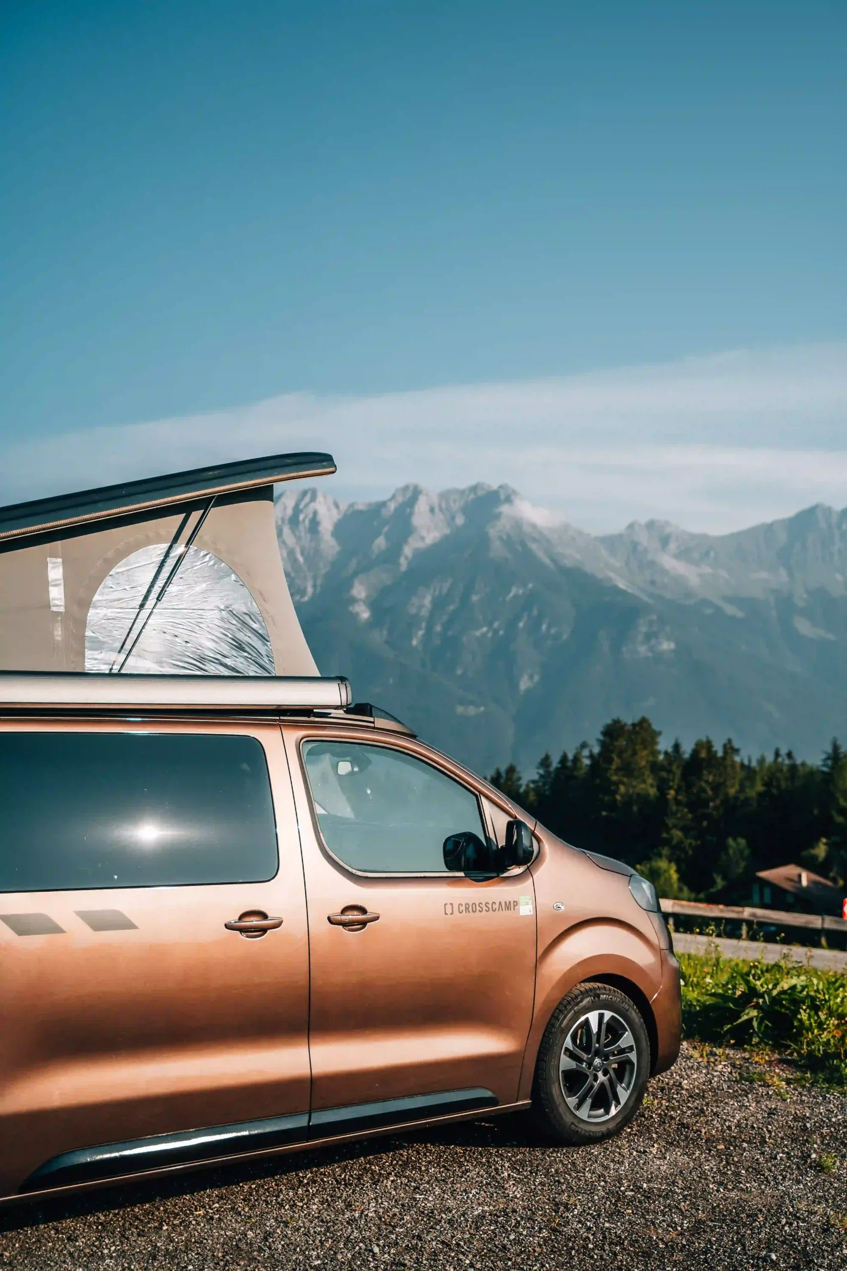 Side view of a bronze campervan with a pop-up roof tent parked in the mountains.
