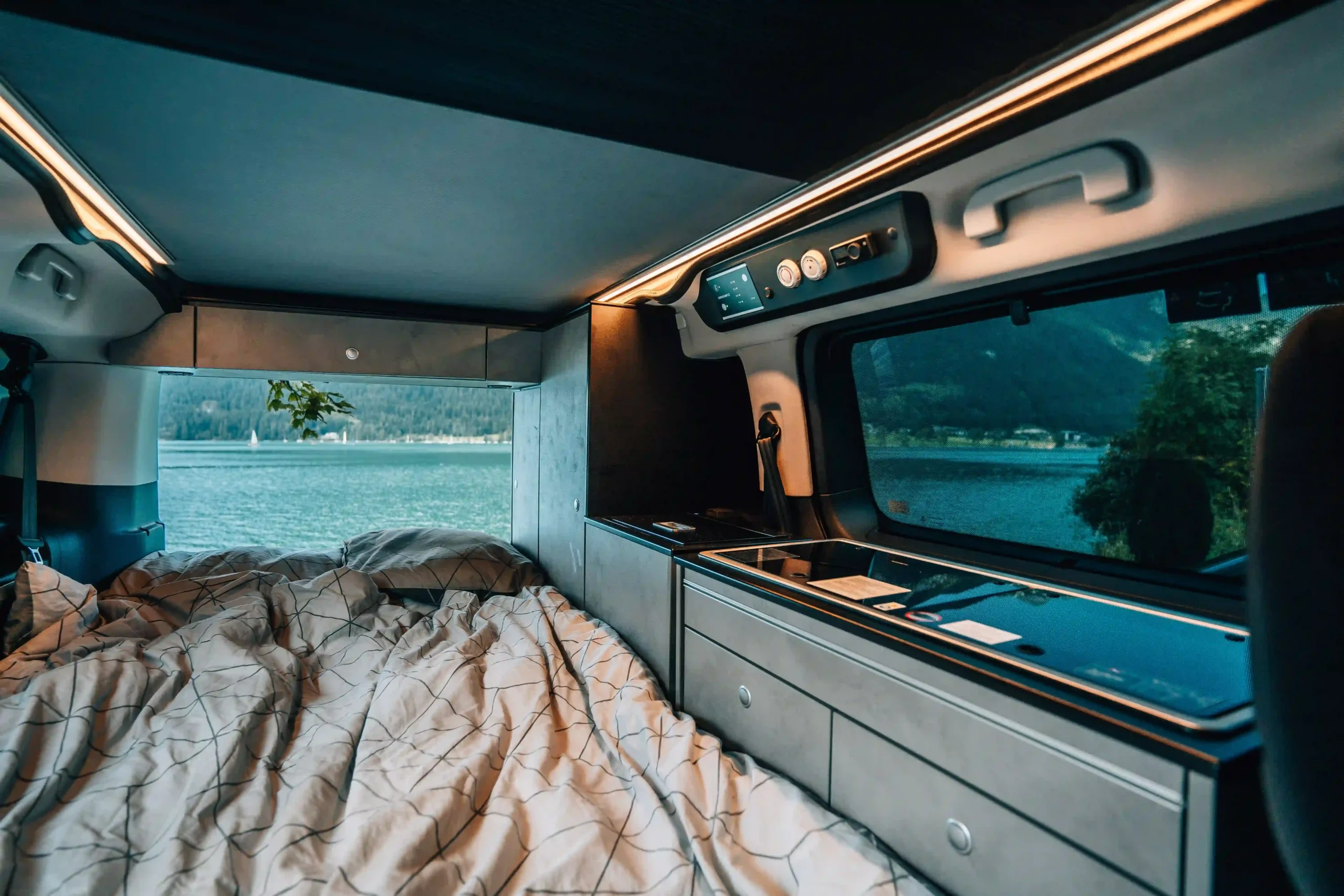 Interior of a campervan showing a bed and kitchenette with a view of a turquoise alpine lake.