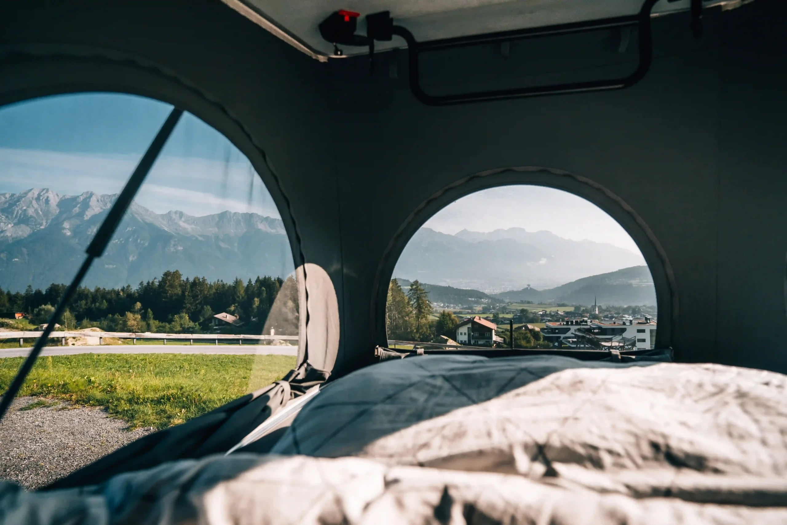 Interior view from the sleeping area of a camper van pop-up tent looking through two rounded windows at a scenic alpine valley.