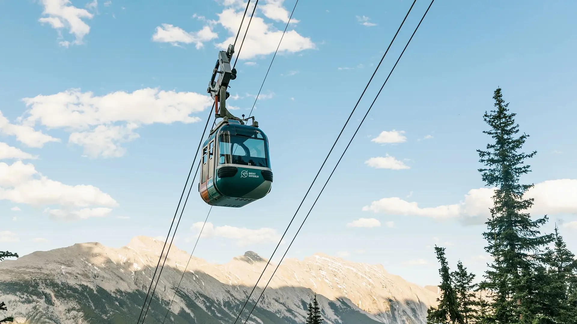 A green and white Banff Gondola cabin suspended on thick cables high above the ground. The cabin is set against a bright blue sky with scattered white clouds, with the rugged, sun-drenched peaks of the Canadian Rockies and tall pine trees visible in the background.