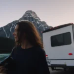 A side profile of a woman holding a camera in front of a large white RV, with the turquoise waters of Lake Louise and a snow-dusted mountain peak in the background.