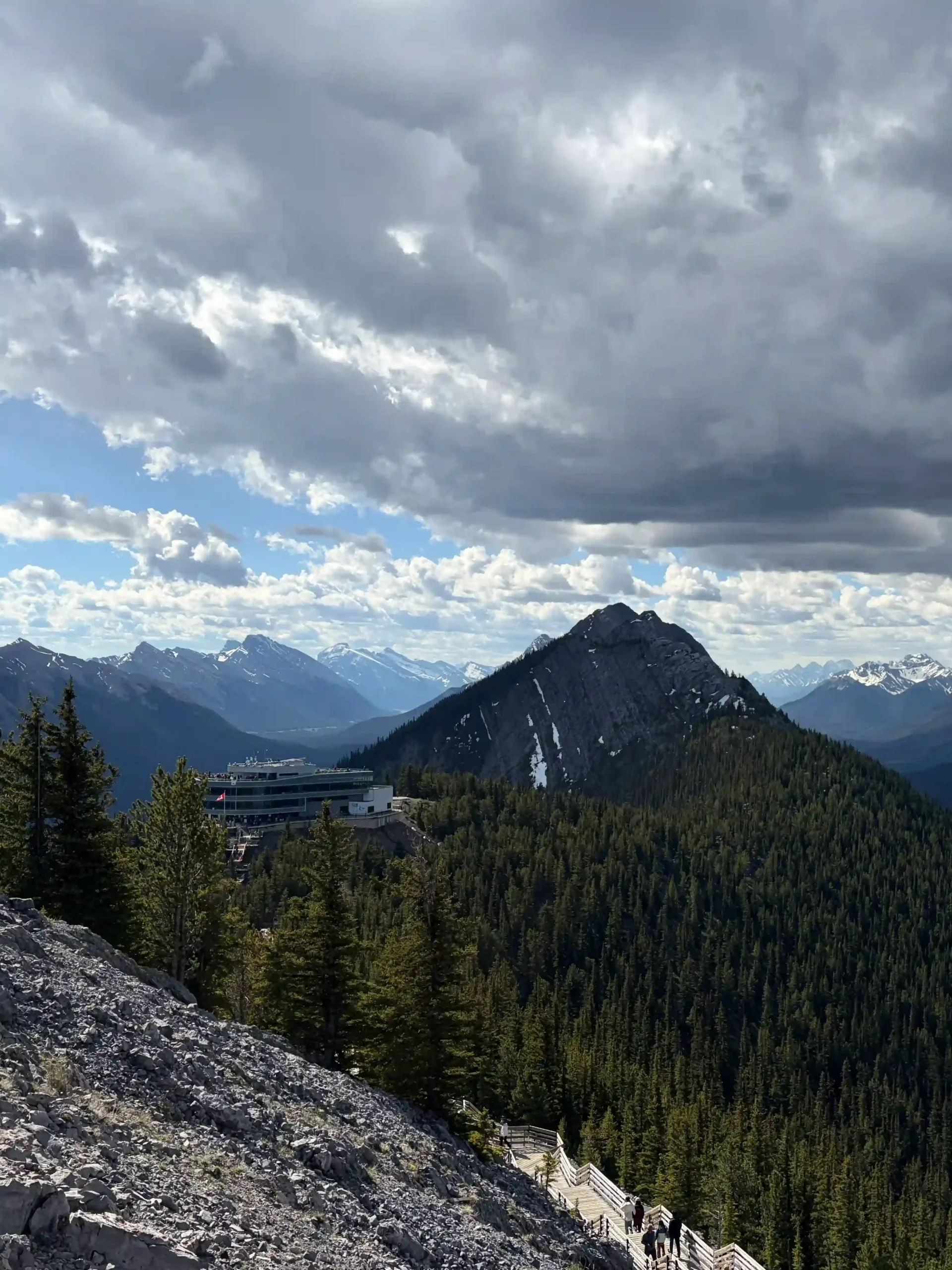 A scenic overlook from a rugged, rocky mountainside showing a wooden boardwalk trail curving down a steep ridge. The modern concrete Banff Gondola upper terminal building sits on a forested cliffside, overlooking a vast pine forest valley.