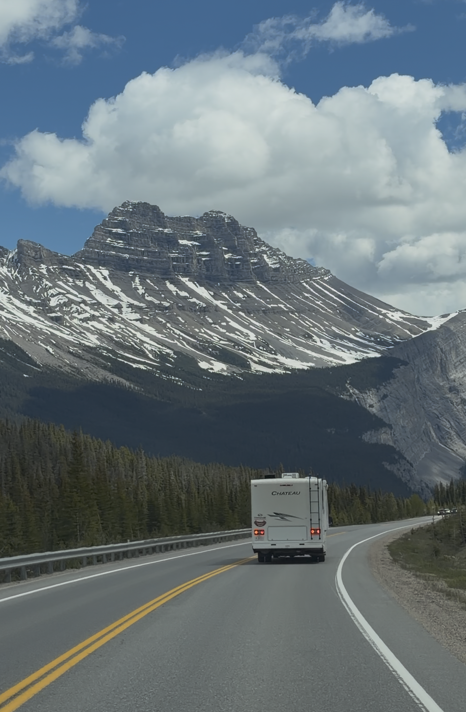 A white motorhome driving along a curved asphalt highway toward a massive snow-dusted mountain peak flanked by pine trees.