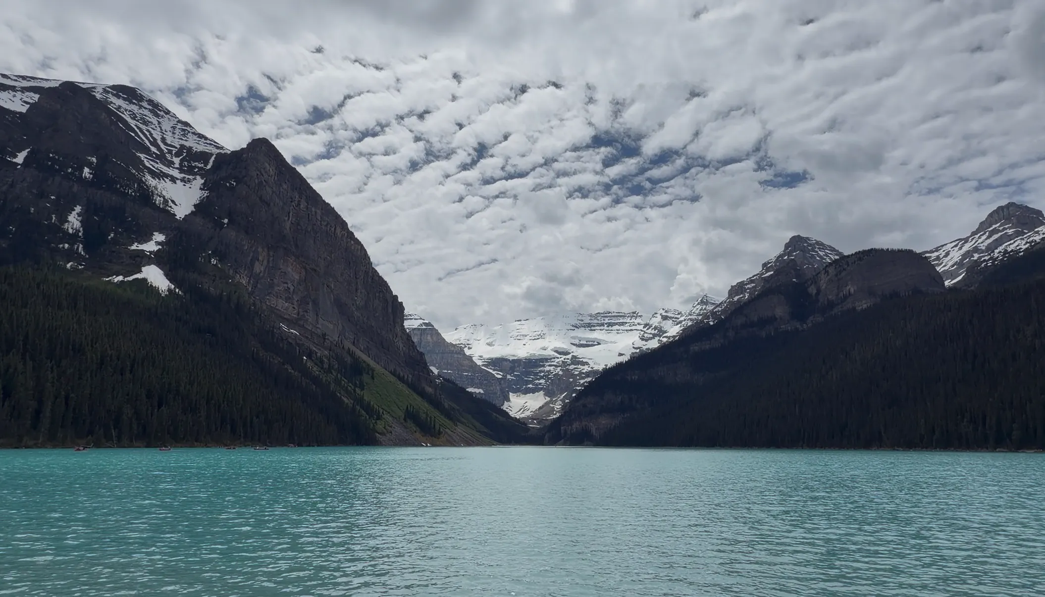 A vibrant turquoise lake flanked by massive dark mountains with a distant snowy glacier under an overcast sky.