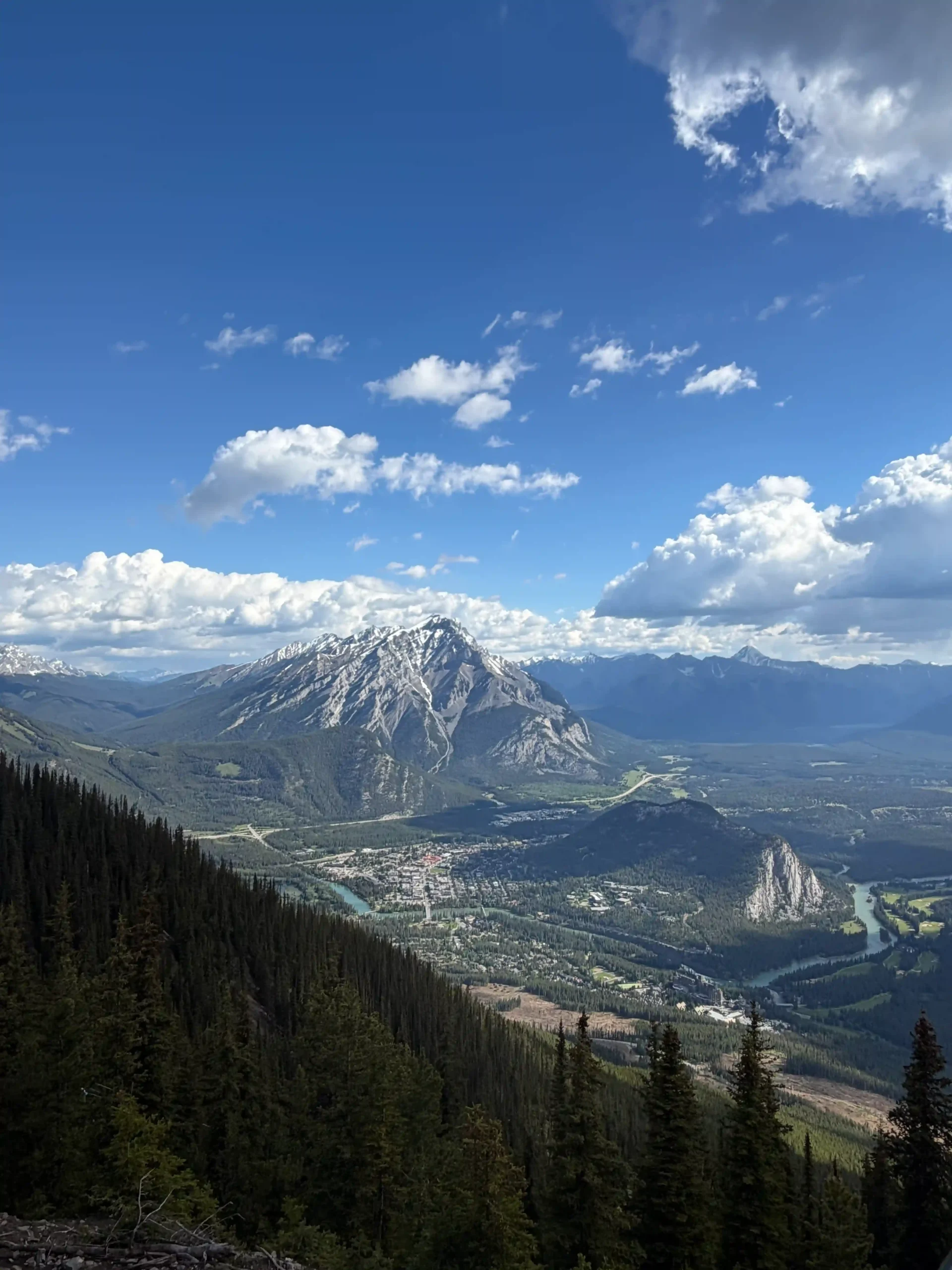 High-angle view of a mountain townsite and a winding blue river surrounded by dense pine forests and rocky peaks.