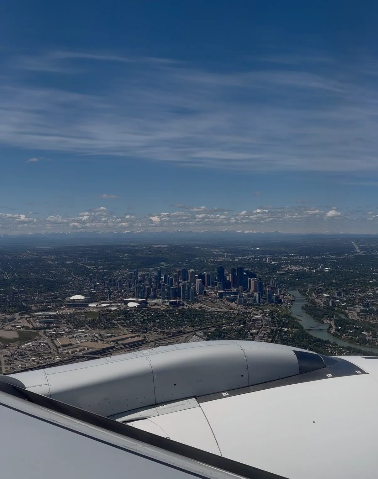 Aerial view of the Calgary downtown skyline and distant mountains from an airplane window over the wing and engine.