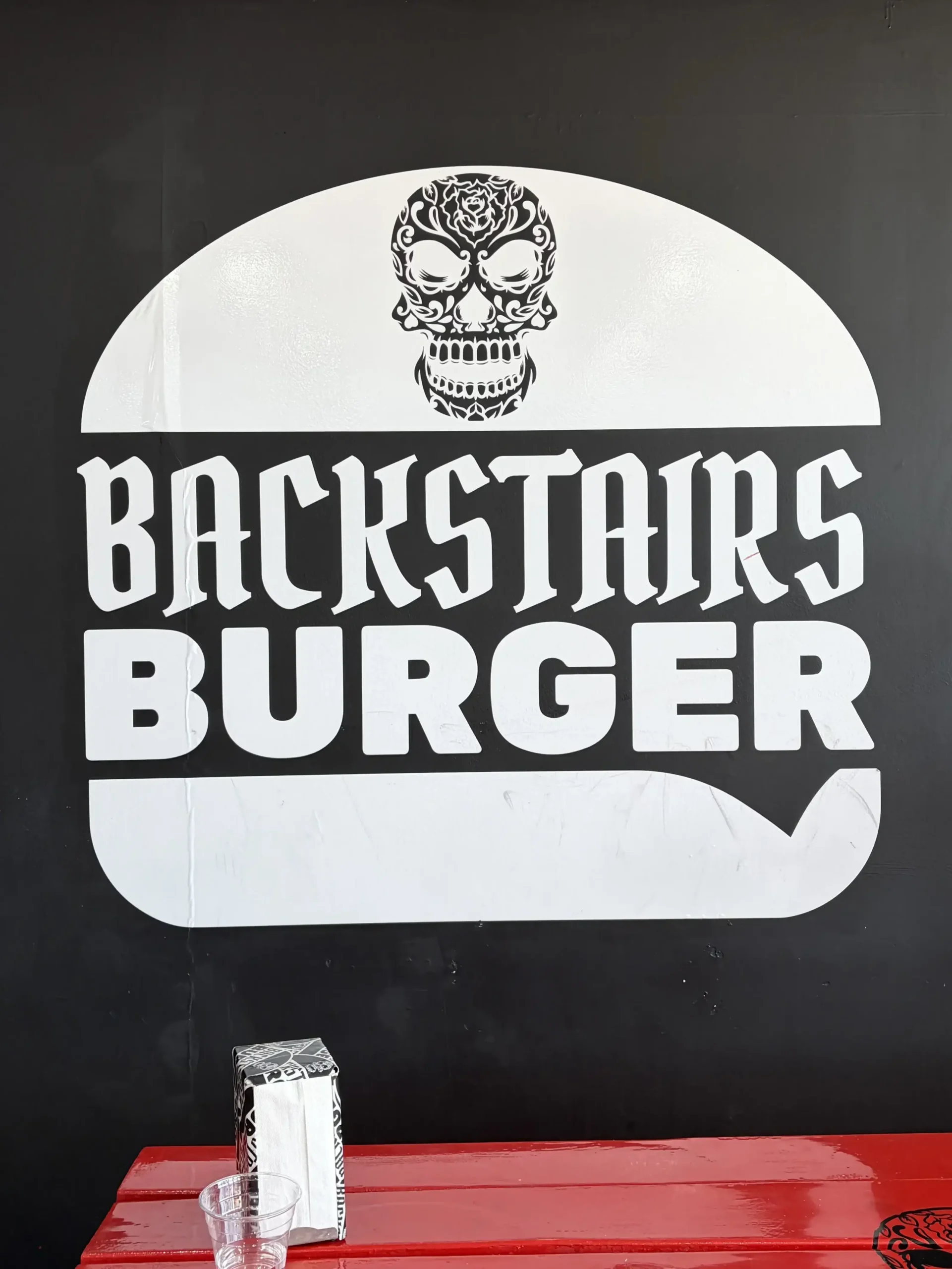A large white logo painted on a black wall, shaped like a hamburger outline. The logo contains a highly detailed skull illustration on top and the stylized gothic text \