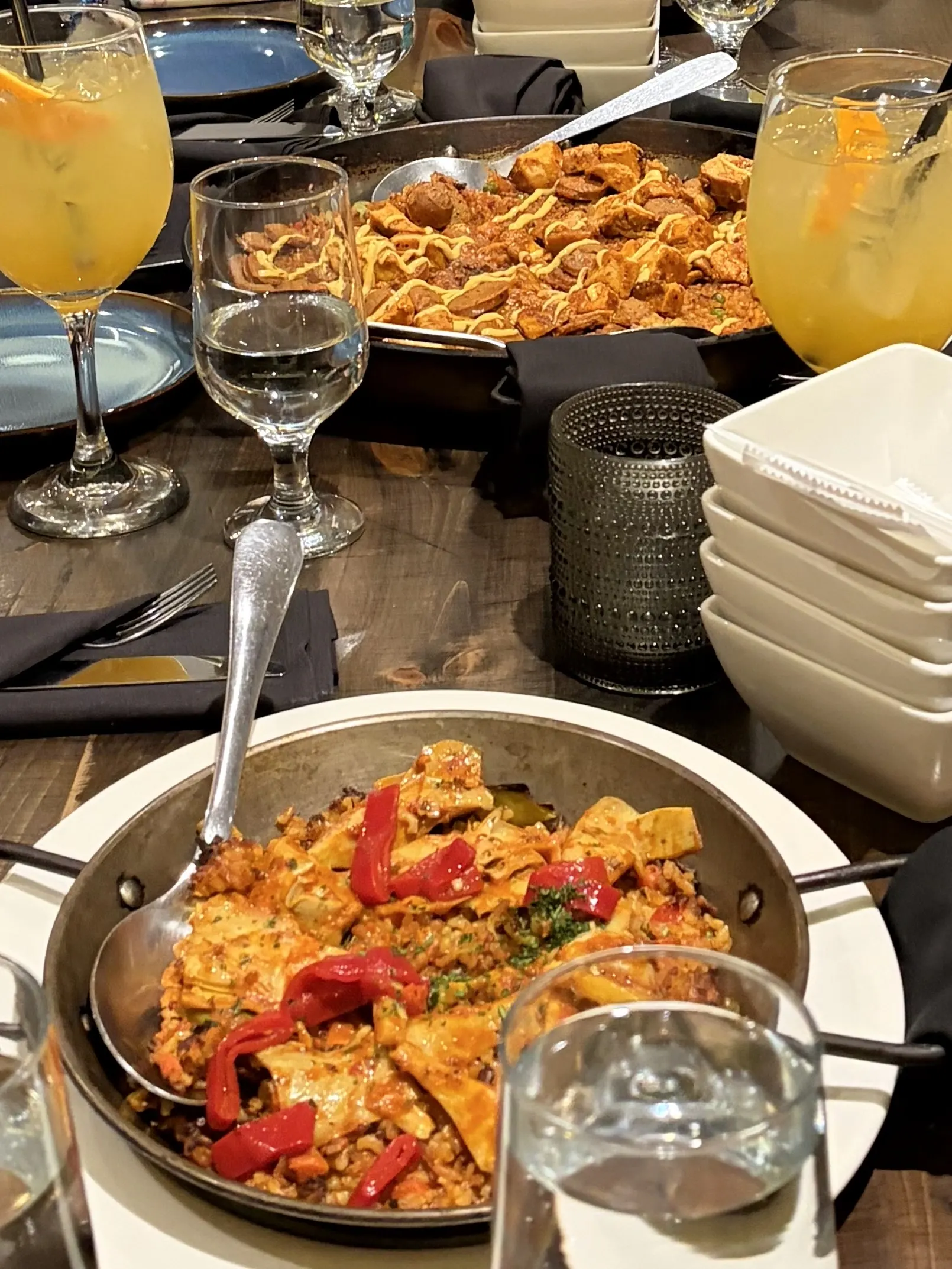 A table filled with Spanish tapas. In the foreground, a small metal pan holds a rice dish topped with sliced red bell peppers. In the background, a larger pan features fried meats. On the table are glasses of yellow sangria with orange slices.